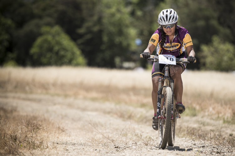A mountain biker in a colorful cycling outfit rides along a dirt path surrounded by dry grass and trees, focused on navigating the terrain. The cyclist wears a helmet and sunglasses for safety while competing in a race.