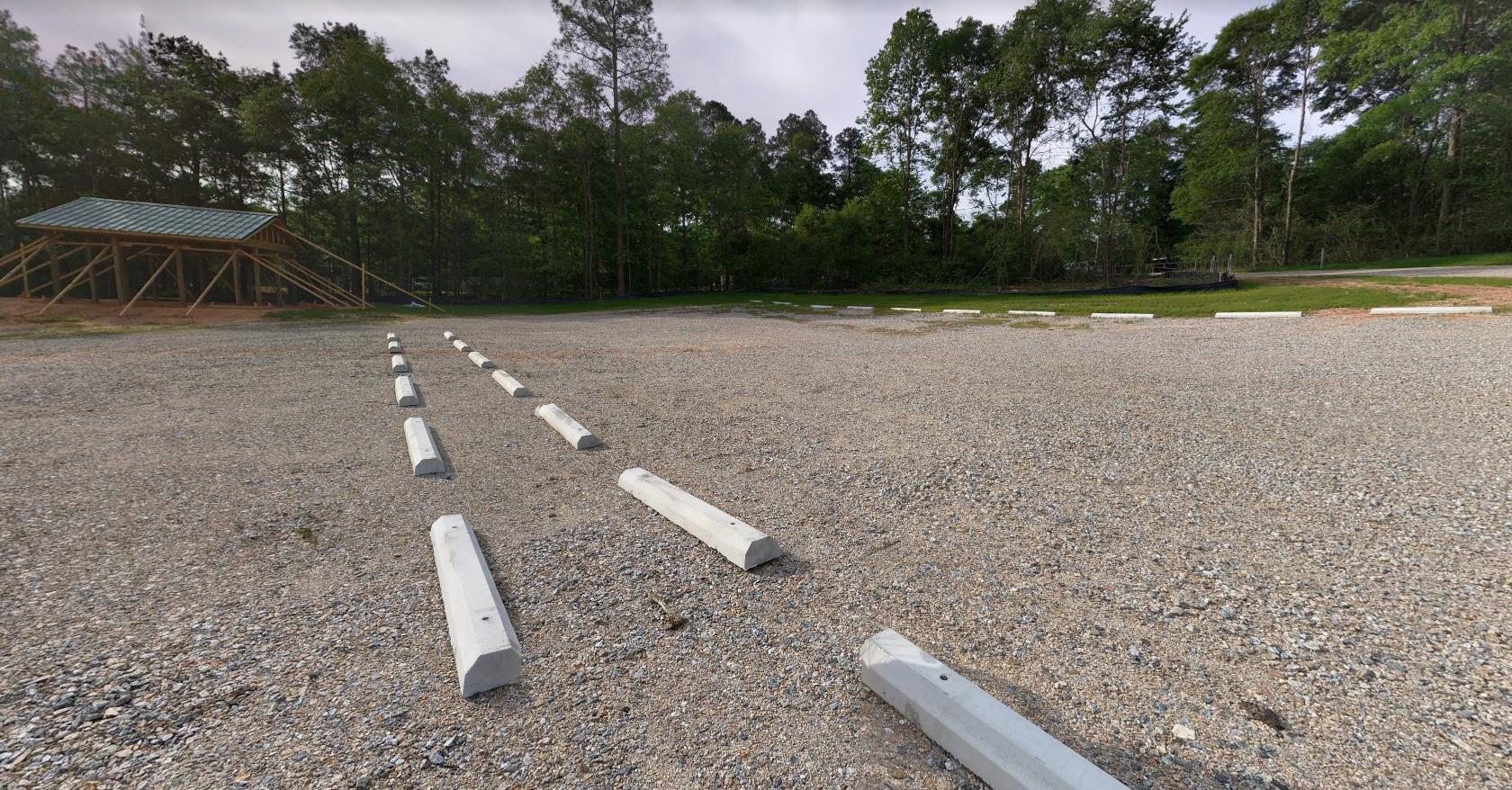 A gravel parking lot with white concrete barriers lined up in rows, set against a backdrop of dense trees. A partially constructed wooden structure with a sloped roof is visible in the background, surrounded by greenery. The area appears quiet and under development. S.A.M.'s Creekside mountain bike trail.