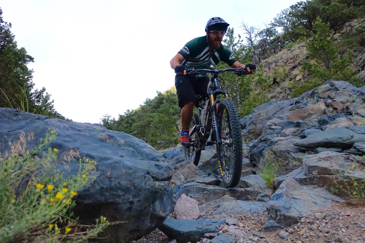 A mountain biker navigating rocky terrain, with lush greenery in the background. The rider is focused and wearing a helmet, gloves, and athletic gear, as they skillfully maneuver over boulders on a trail. Cottonwood mountain bike trail.