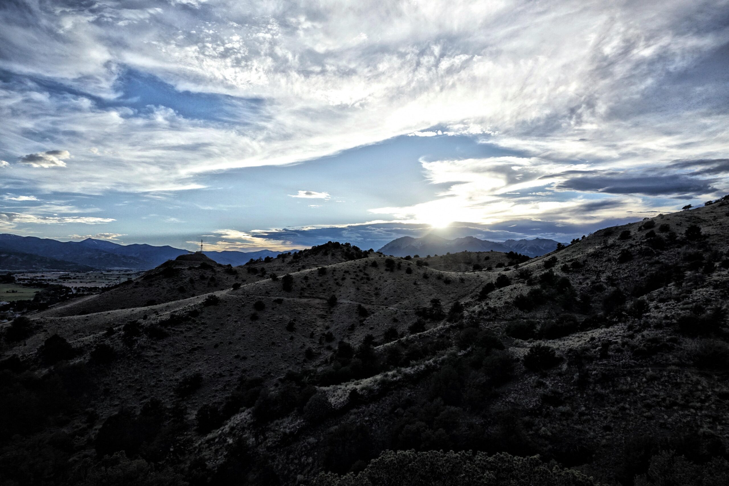 A scenic landscape featuring rolling hills silhouetted against a sunset. The sky is filled with clouds in varying shades of blue and white, while the sun sets behind distant mountains, casting a warm glow. Sparse vegetation punctuates the hilly terrain, creating a peaceful and picturesque natural scene. Cottonwood mountain bike trail.