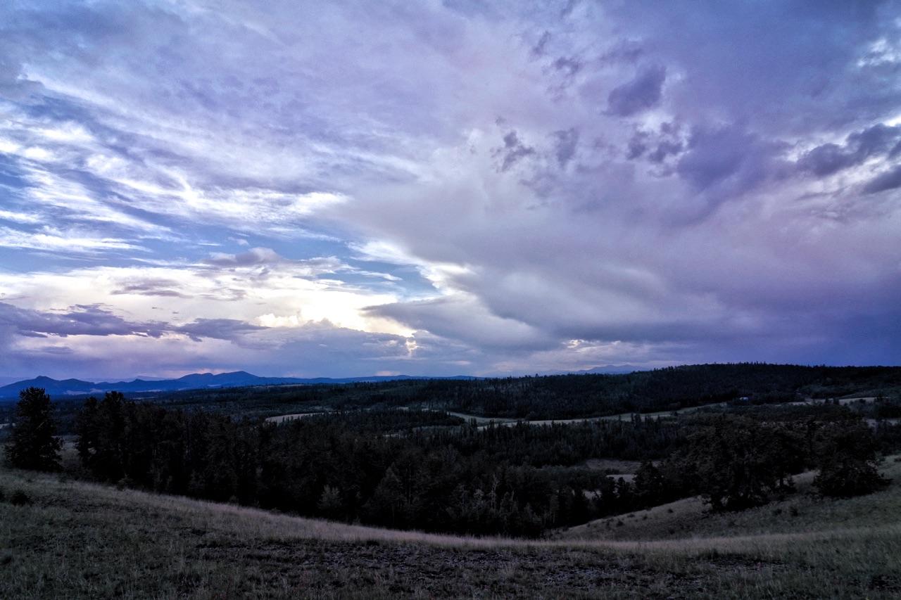 A panoramic view of a landscape at dusk, featuring rolling hills, dense forests, and distant mountains under a dramatic sky filled with clouds in shades of blue and purple. The scene conveys a serene and tranquil atmosphere. Colorado Trail: Kenosha Pass To Lost Creek Wilderness Bound mountain bike trail.