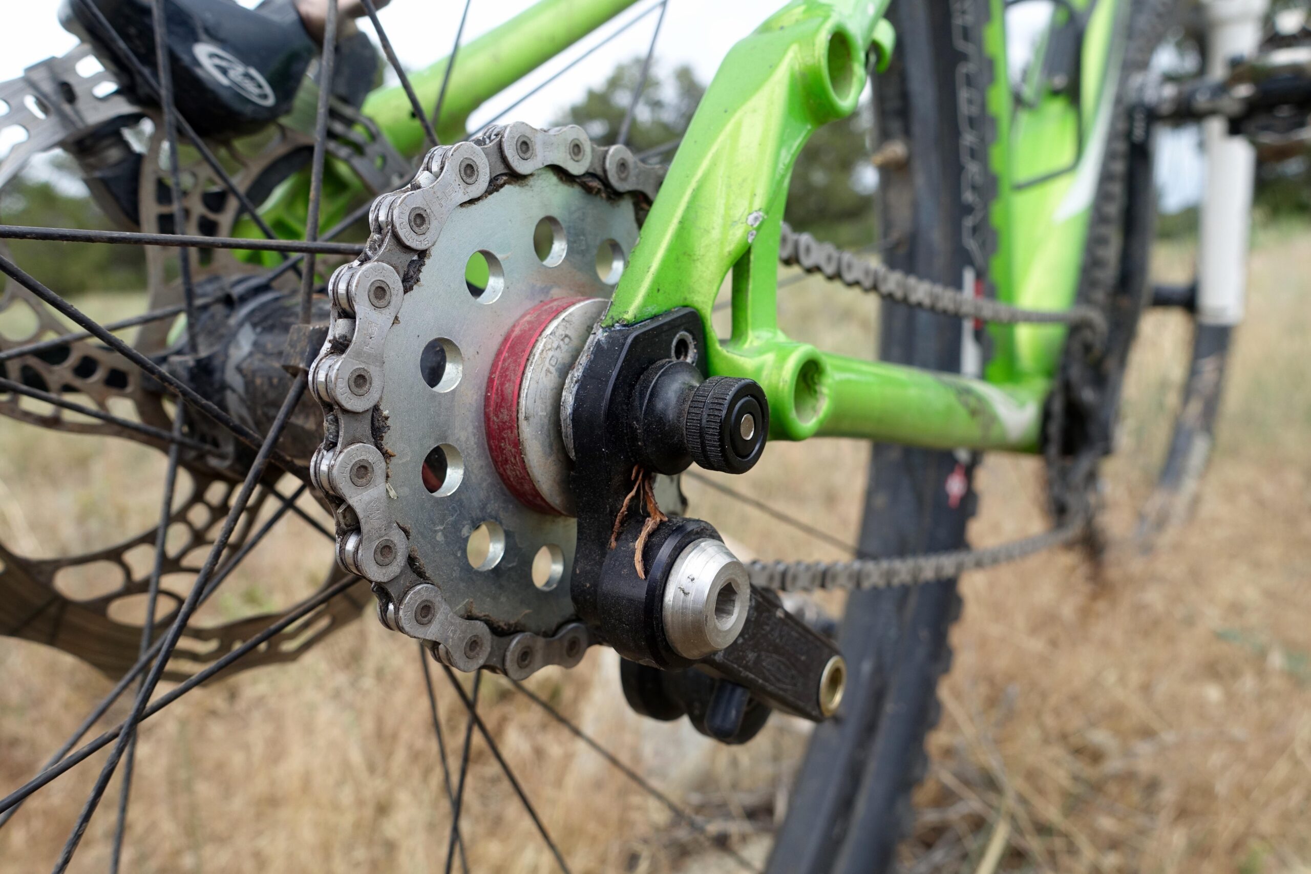 Airborne Goblin: Close-up view of a bicycle's rear drivetrain, showcasing the chain, sprocket, and derailleur against a natural background of dry grass. The bike frame is green, with intricate details of the components highlighted.