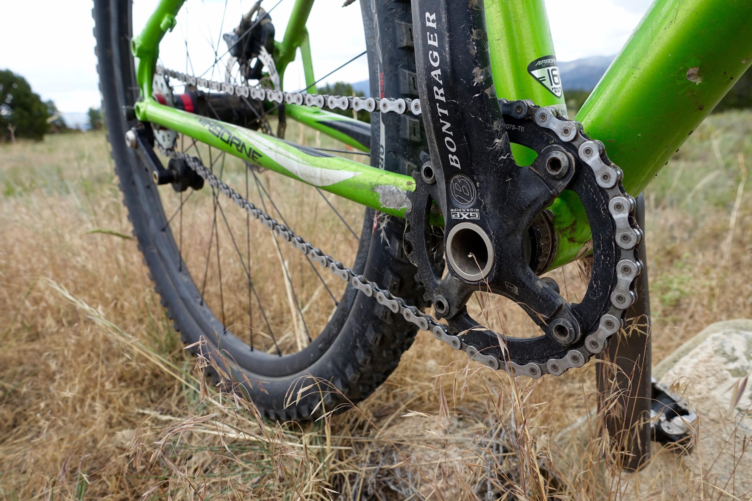 Airborne Goblin: Close-up of a green mountain bike's drivetrain, showcasing the chain, crankset, and rear wheel. The bike is positioned in a grassy outdoor setting.