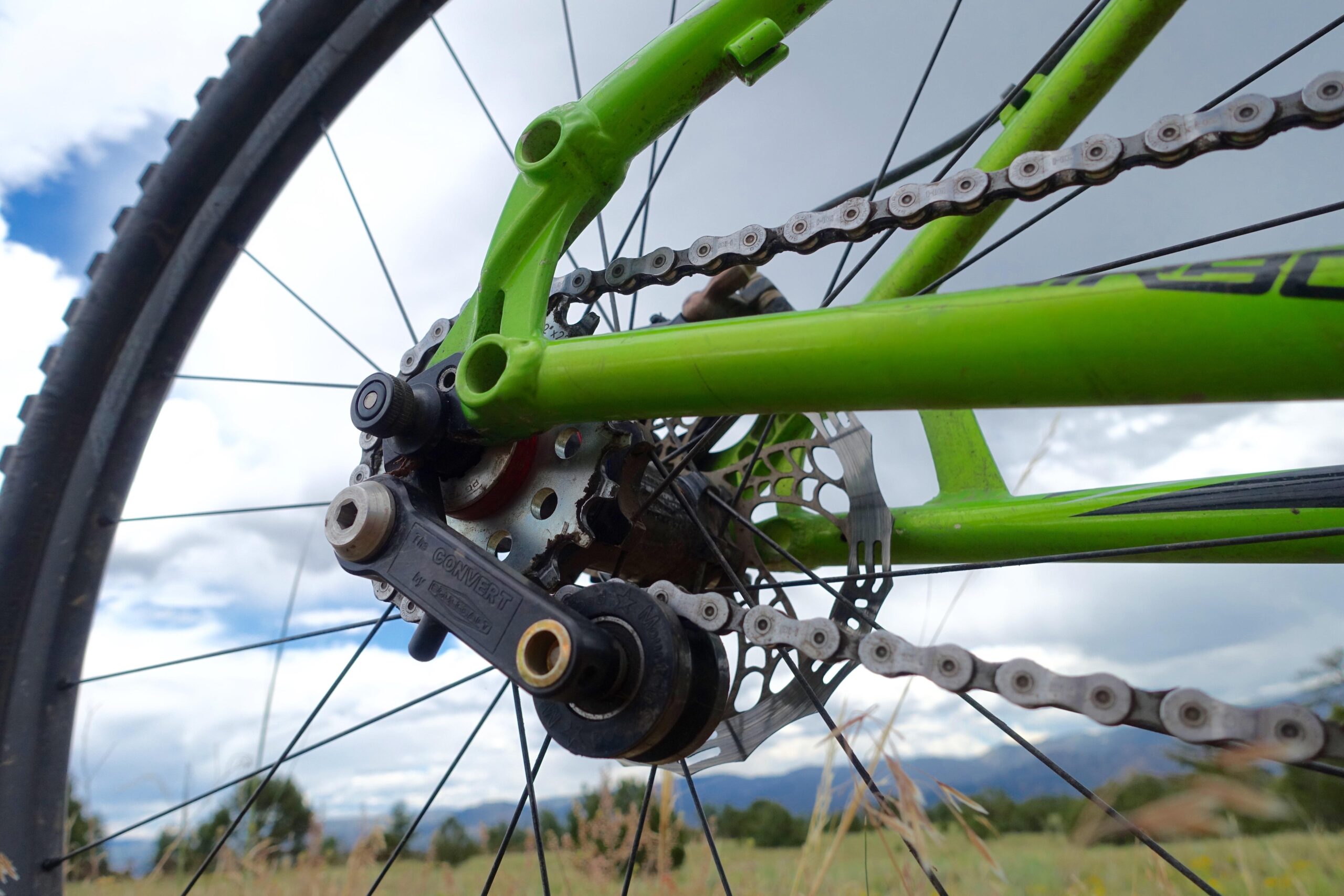 Airborne Goblin: Close-up view of the rear derailleur and chain of a green mountain bike, set against a backdrop of blue skies and distant mountains. The bike's frame, chain, and cassette are prominently featured, highlighting the bike's mechanical components and the outdoor environment.
