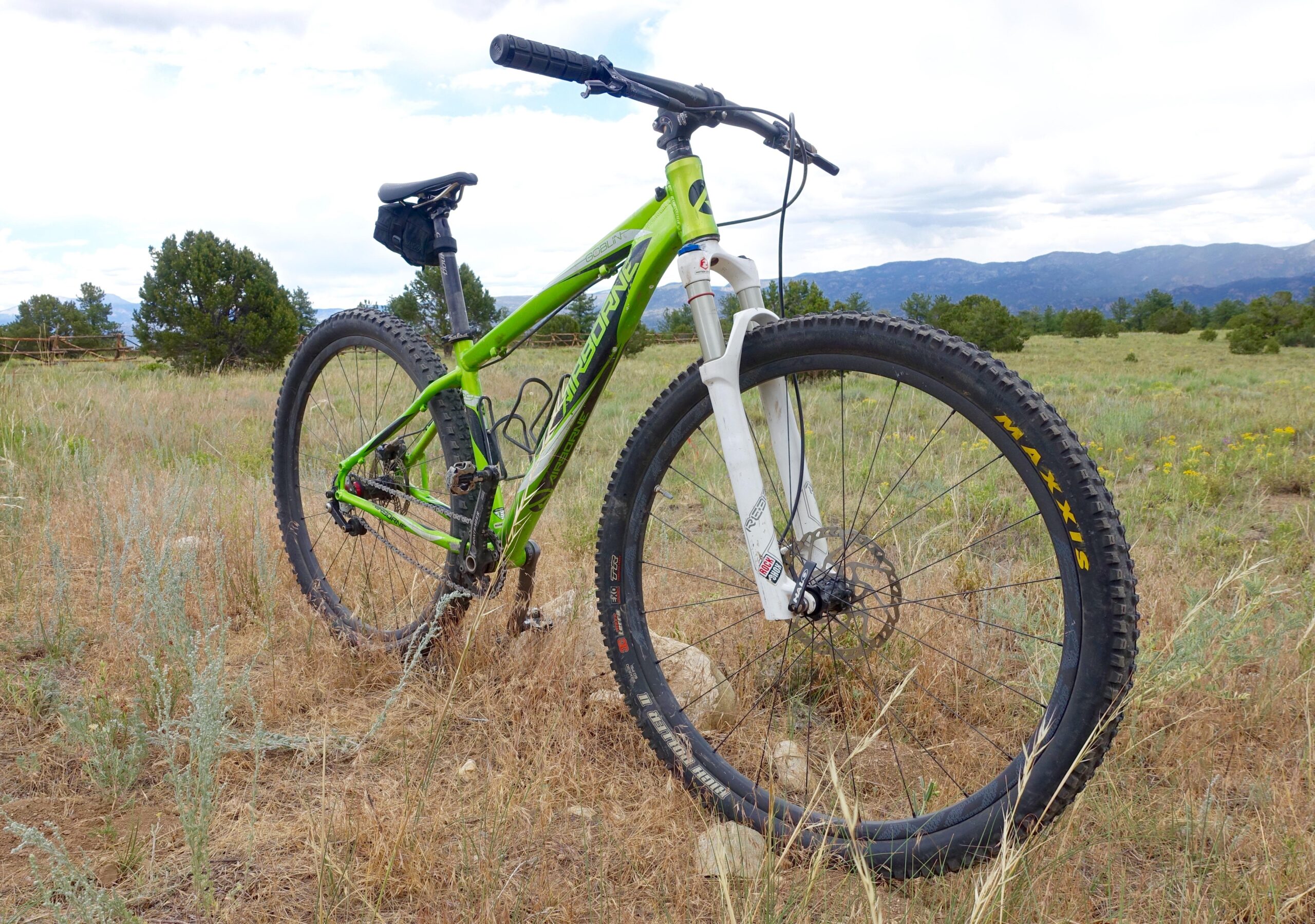 Airborne Goblin: A green mountain bike with a white front fork is parked in an open field, surrounded by tall grass and shrubs, with mountains in the background under a cloudy sky.