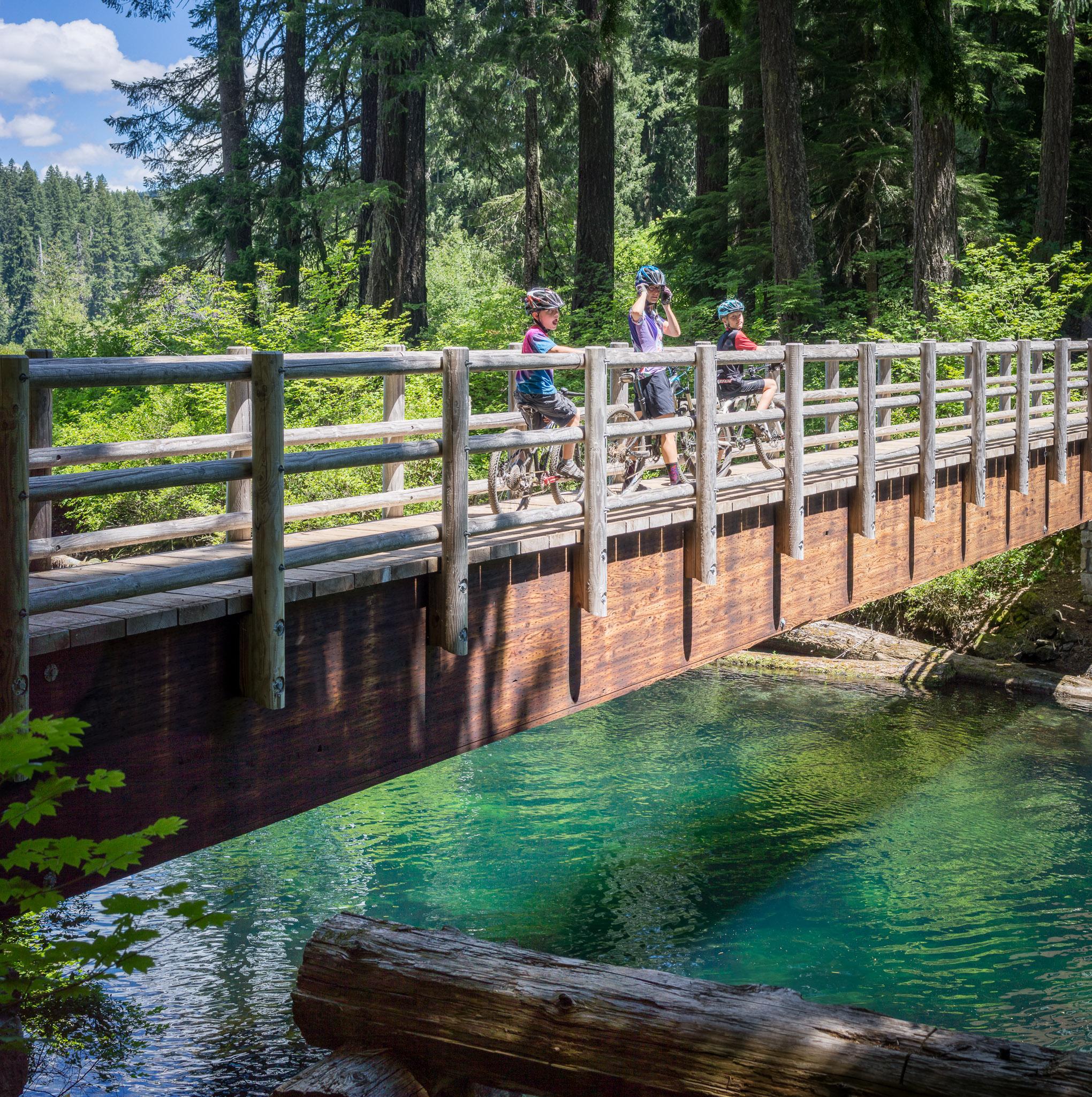 Three cyclists riding their bikes on a wooden bridge over a clear, turquoise stream. Surrounded by lush green trees and under a bright sky with scattered clouds, the scene captures a vibrant outdoor setting perfect for biking and nature exploration. Mckenzie River Trail mountain bike trail.