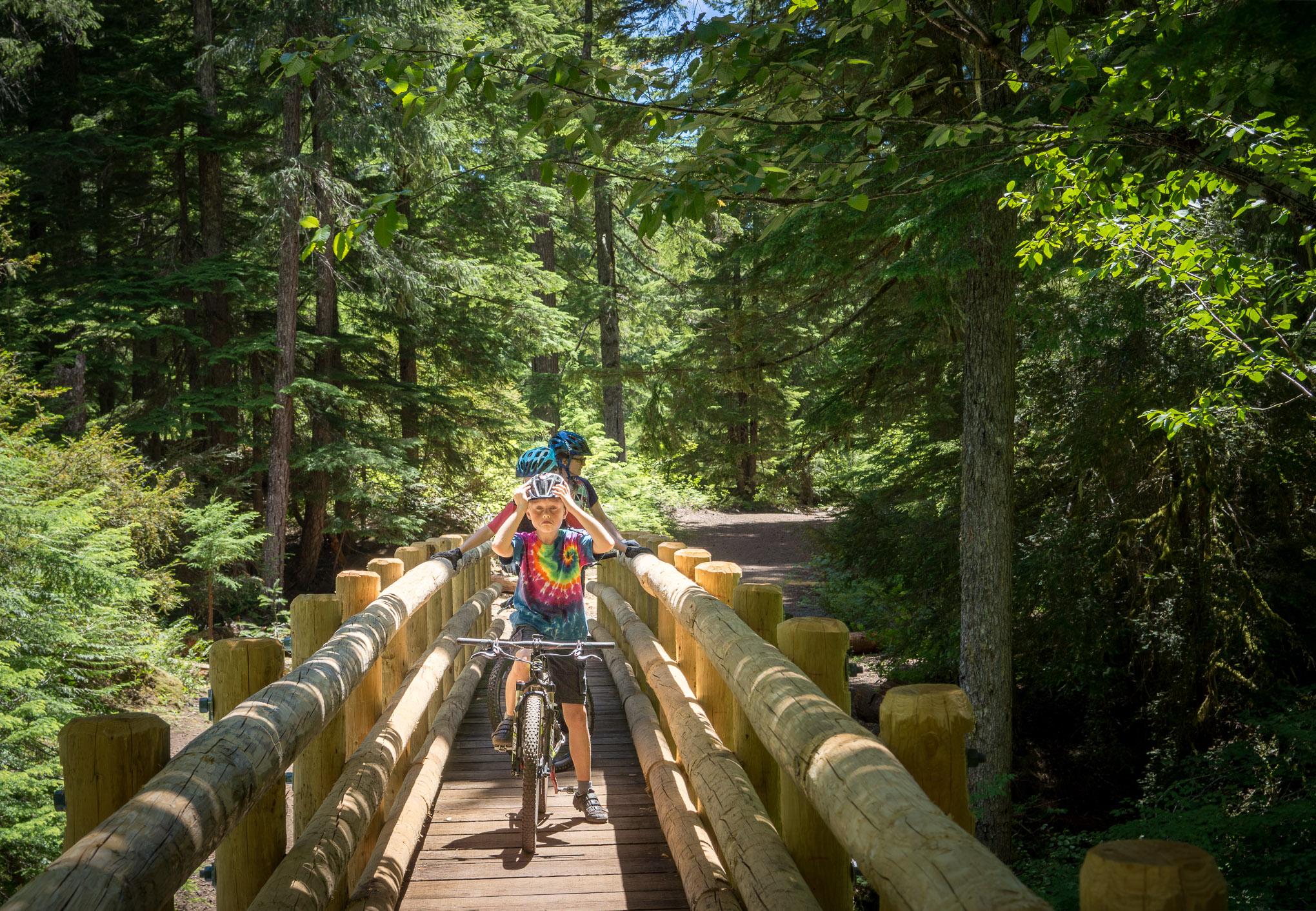 A child in a colorful tie-dye shirt and a helmet is riding a bicycle across a wooden bridge in a lush forest. The scene is filled with green trees and sunlight filtering through the leaves, creating a vibrant and playful atmosphere. Another child can be seen in the background, also wearing a helmet. Mckenzie River Trail mountain bike trail.
