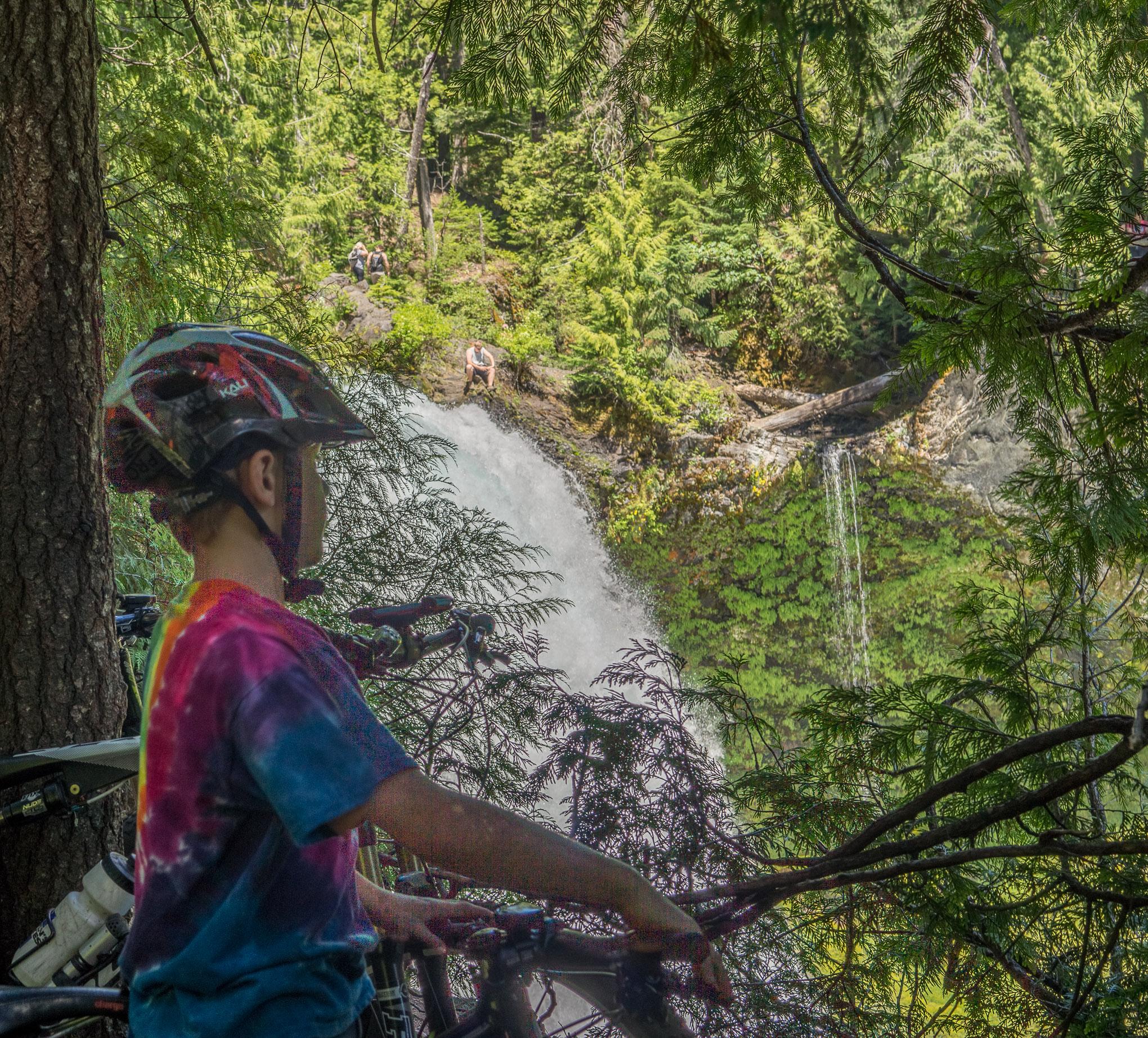A child wearing a colorful tie-dye shirt and a cycling helmet stands beside a mountain bike, gazing at a waterfall surrounded by lush greenery. In the background, two people are seen sitting on rocks near the waterfall, enjoying the natural scenery. Mckenzie River Trail mountain bike trail.
