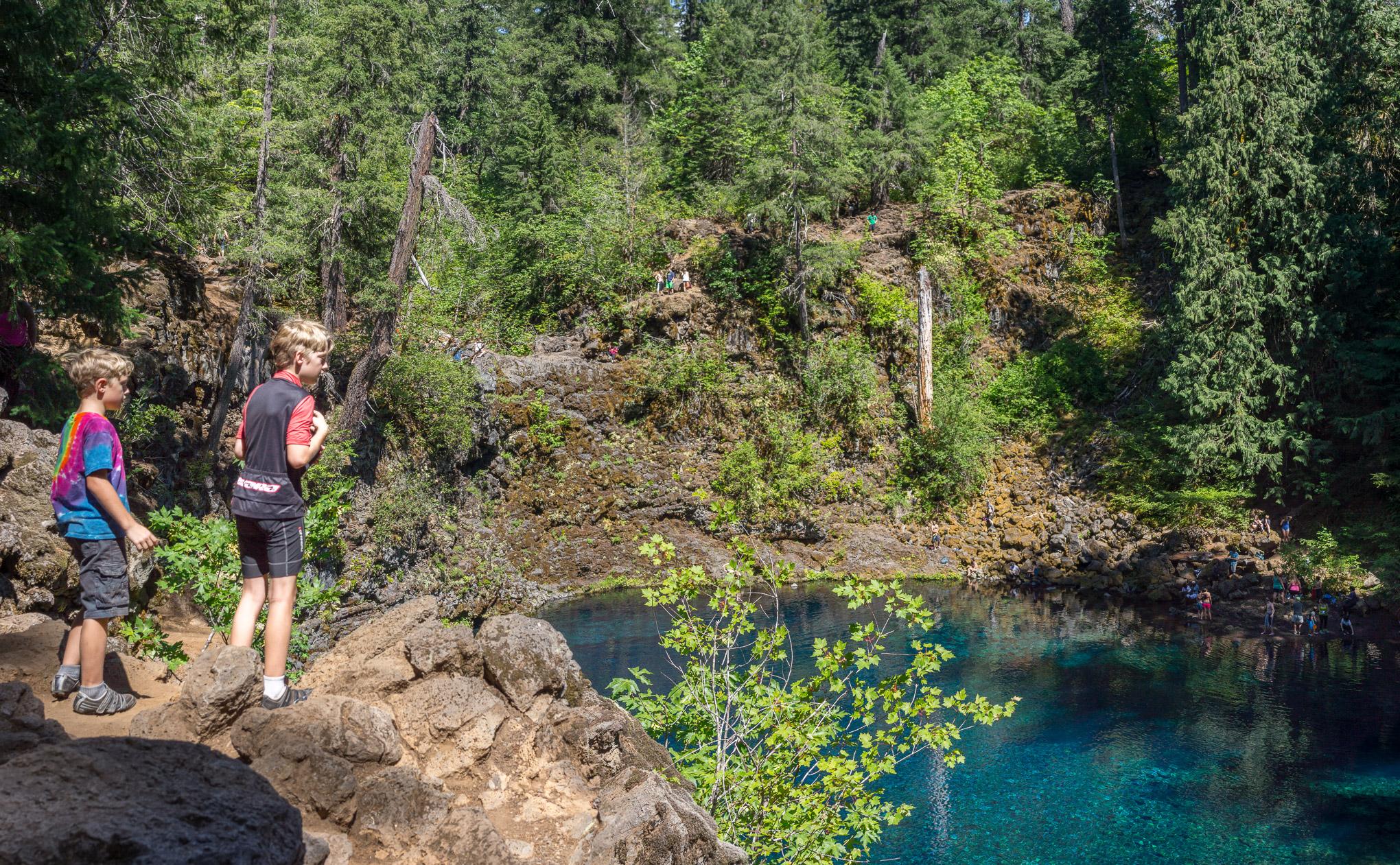 Two children stand on rocky terrain near a clear blue body of water, surrounded by lush green trees. One child wears a colorful tie-dye shirt while the other is dressed in a dark vest and shorts. In the background, several people are seen on the rocky shore and at the water's edge, enjoying the scenic outdoors. The sunlight filters through the trees, creating a vibrant and inviting atmosphere. Mckenzie River Trail mountain bike trail.