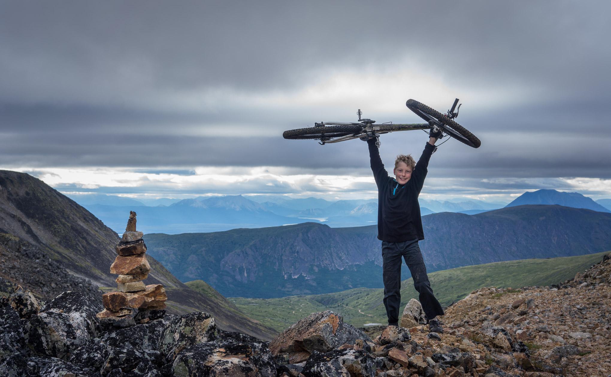 A child joyfully holds a mountain bike above their head while standing on rocky terrain at a high elevation, with mountain ranges and cloudy skies in the background. A small stack of rocks is visible nearby. Atlin Mt SuperHero Trail mountain bike trail.