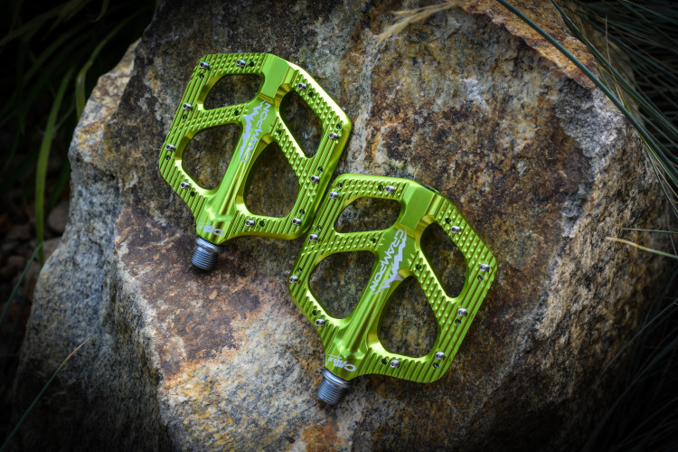 Bright green bicycle pedals are positioned on a textured rock surface, showcasing their intricate design and metal texture. The pedals feature multiple grip pins for added traction and have a sleek, modern appearance. Surrounding foliage partially frames the image, highlighting the outdoor setting.