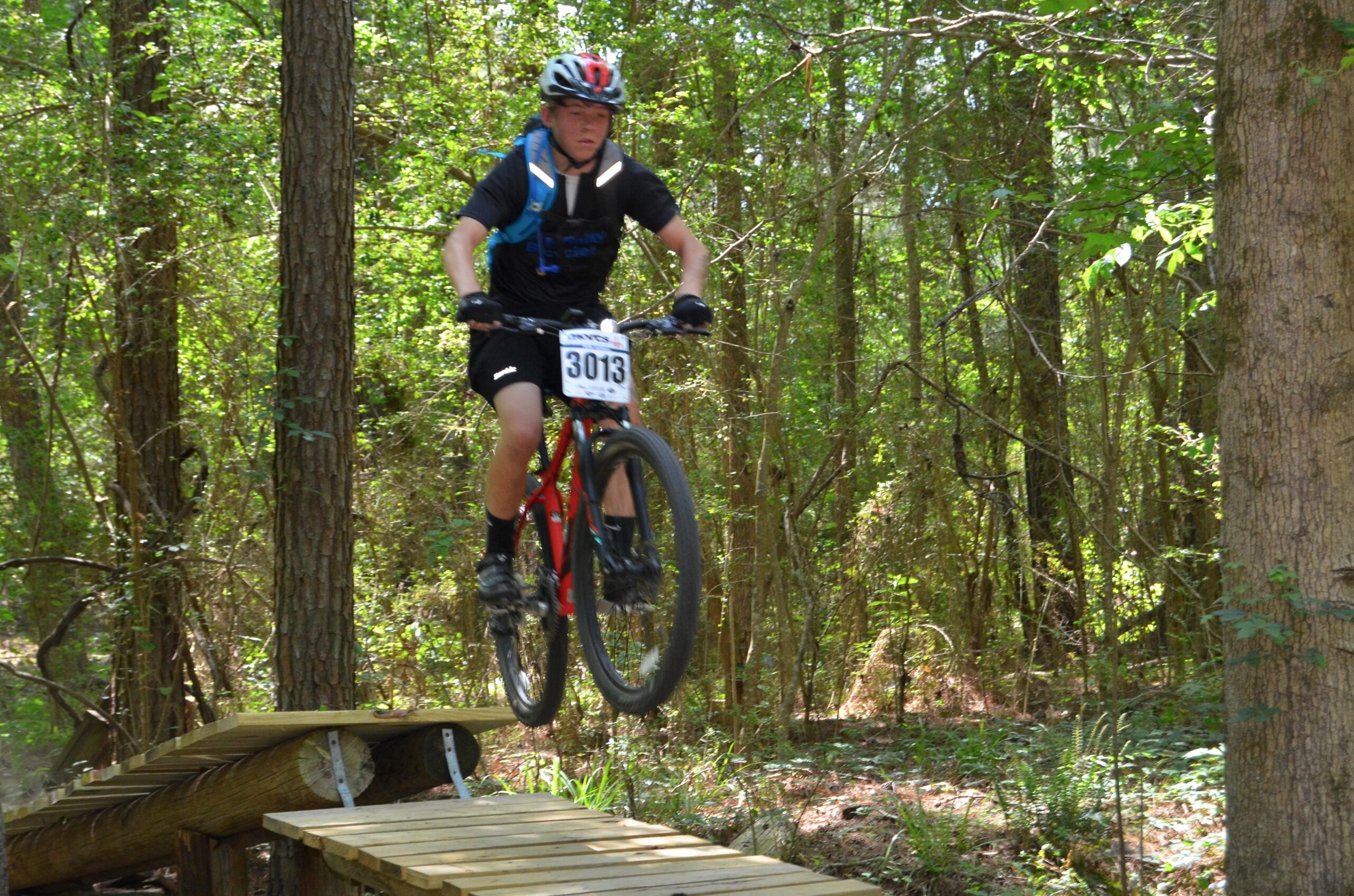 A young cyclist in a black helmet and blue backpack is airborne while jumping off a wooden ramp in a forested area. He is wearing a black shirt and shorts, with a race number visible on his shirt. Surrounding him are tall trees and lush greenery. Mt. Zion Bike Trails mountain bike trail.