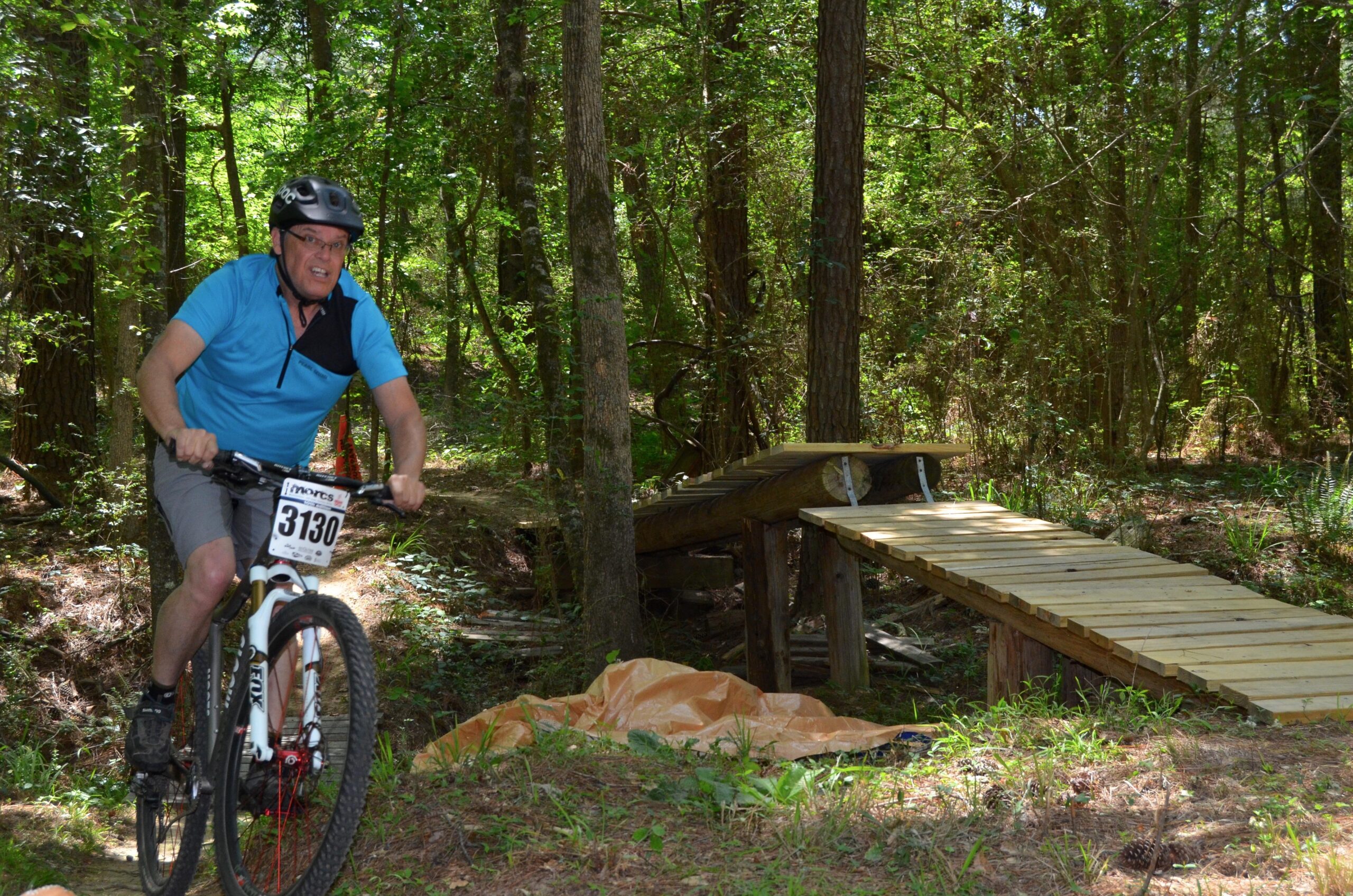 A man in a blue shirt riding a mountain bike on a trail in a wooded area, approaching a wooden bridge. The scene is surrounded by vibrant greenery, with trees and underbrush in the background. The rider is wearing a helmet and is focused on navigating the terrain. The ground is covered with grass and patches of sunlight filter through the trees. Mt. Zion Bike Trails mountain bike trail.