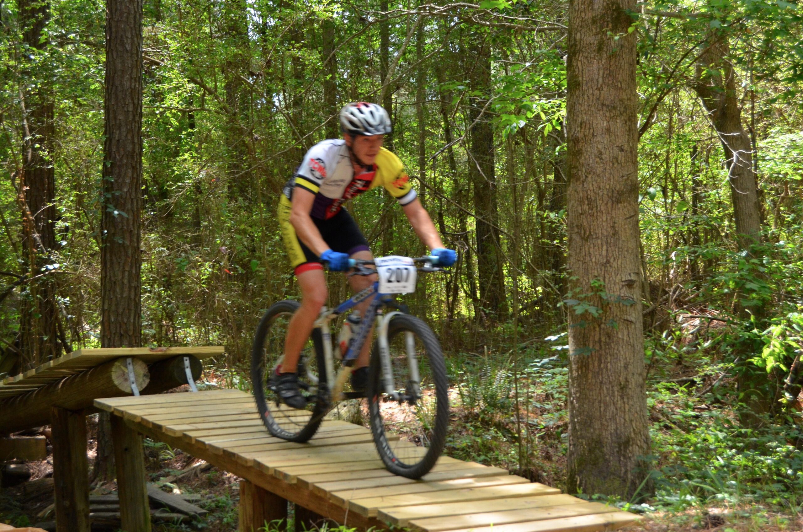 A mountain biker in a colorful racing outfit is riding over a wooden bridge on a forest trail, surrounded by lush greenery. The cyclist is focused and in motion, showcasing a competitive spirit in the outdoor setting. The background features tall trees and dense foliage, emphasizing the natural environment of the race. Mt. Zion Bike Trails mountain bike trail.