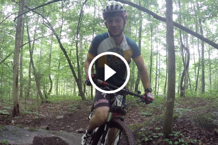 A person on a mountain bike smiles while standing on a wooded trail, surrounded by lush green trees and underbrush. The setting suggests a recreational outdoor environment for cycling. A play button overlay indicates the image is part of a video.
