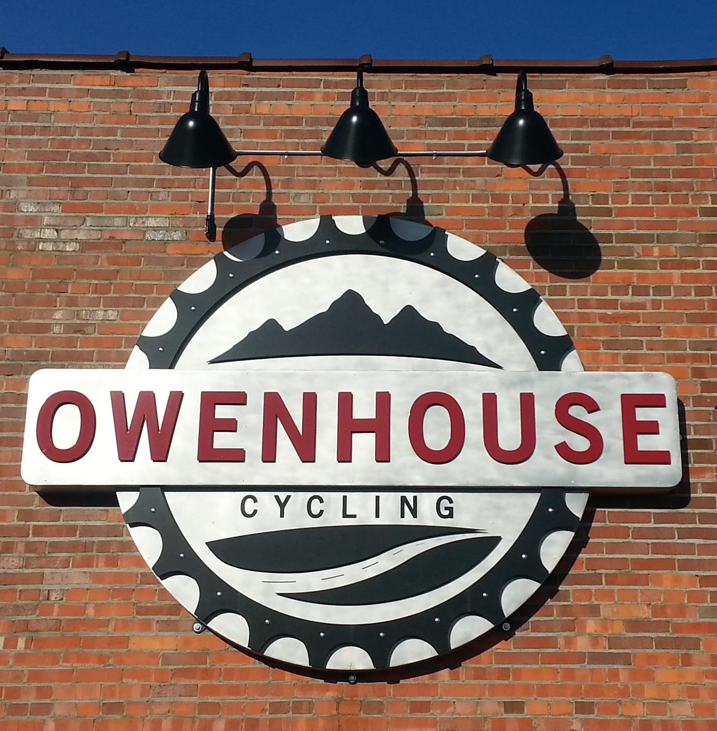 Sign for Owenhouse Cycling, featuring a circular design with mountains and a bicycle gear motif, set against a brick wall. The name "OWENHOUSE" is prominently displayed in red, with "CYCLING" in black below it. Two black light fixtures are positioned above the sign.