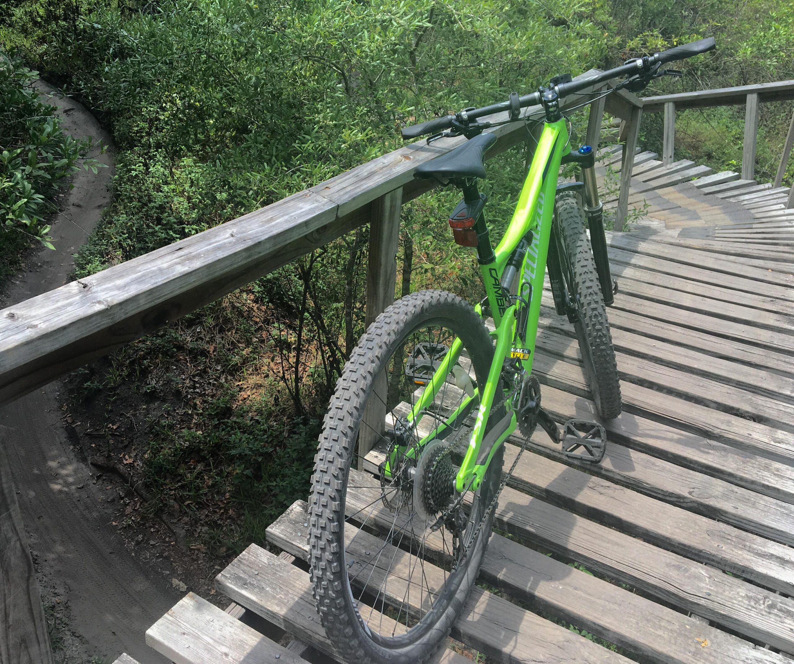 A bright green mountain bike rests on a wooden bridge surrounded by lush greenery, with a winding dirt path visible in the background. Markham Park mountain bike trail.