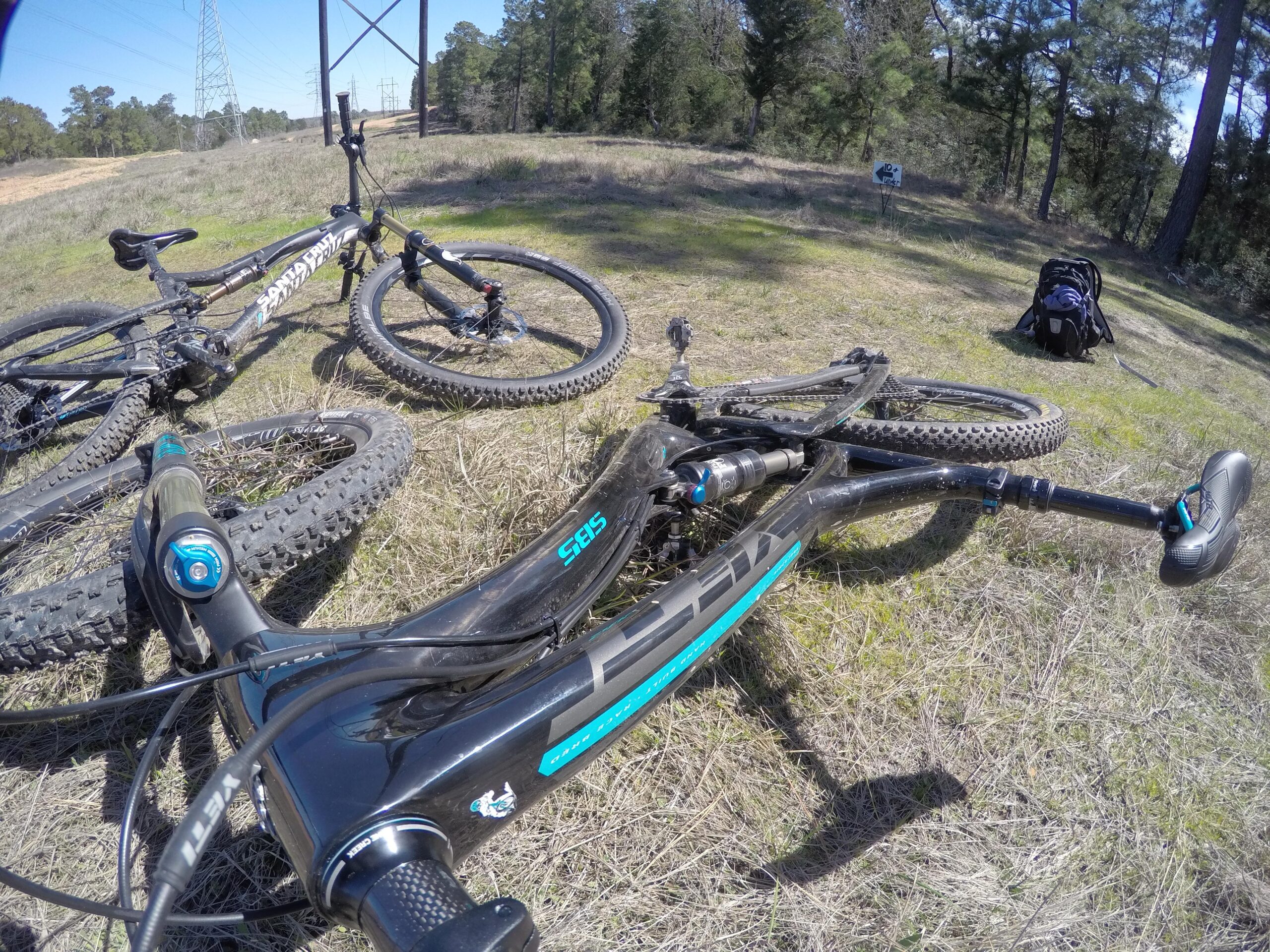 Yeti SB5 Enduro: A close-up view of a mountain bike lying on a grassy area, featuring a black and blue frame with the brand name visible. Another mountain bike is positioned nearby, with a backpack in the background and trees surrounding the scene. The image captures a tranquil outdoor setting, ideal for cycling enthusiasts.