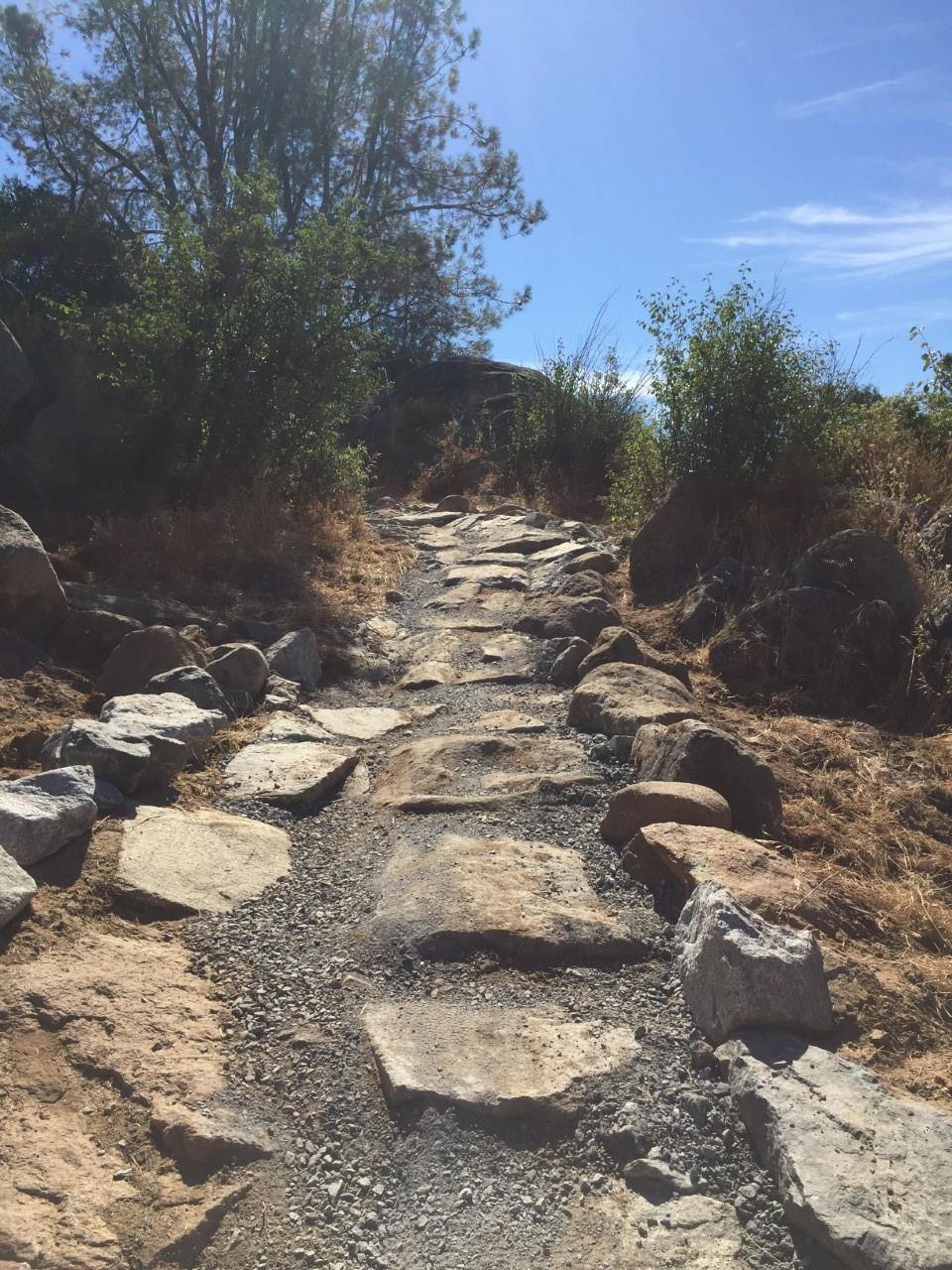 A rugged hiking path made of large stones and gravel, lined with sparse greenery and surrounded by rocky terrain under a clear blue sky. Granite Bay Trail mountain bike trail.