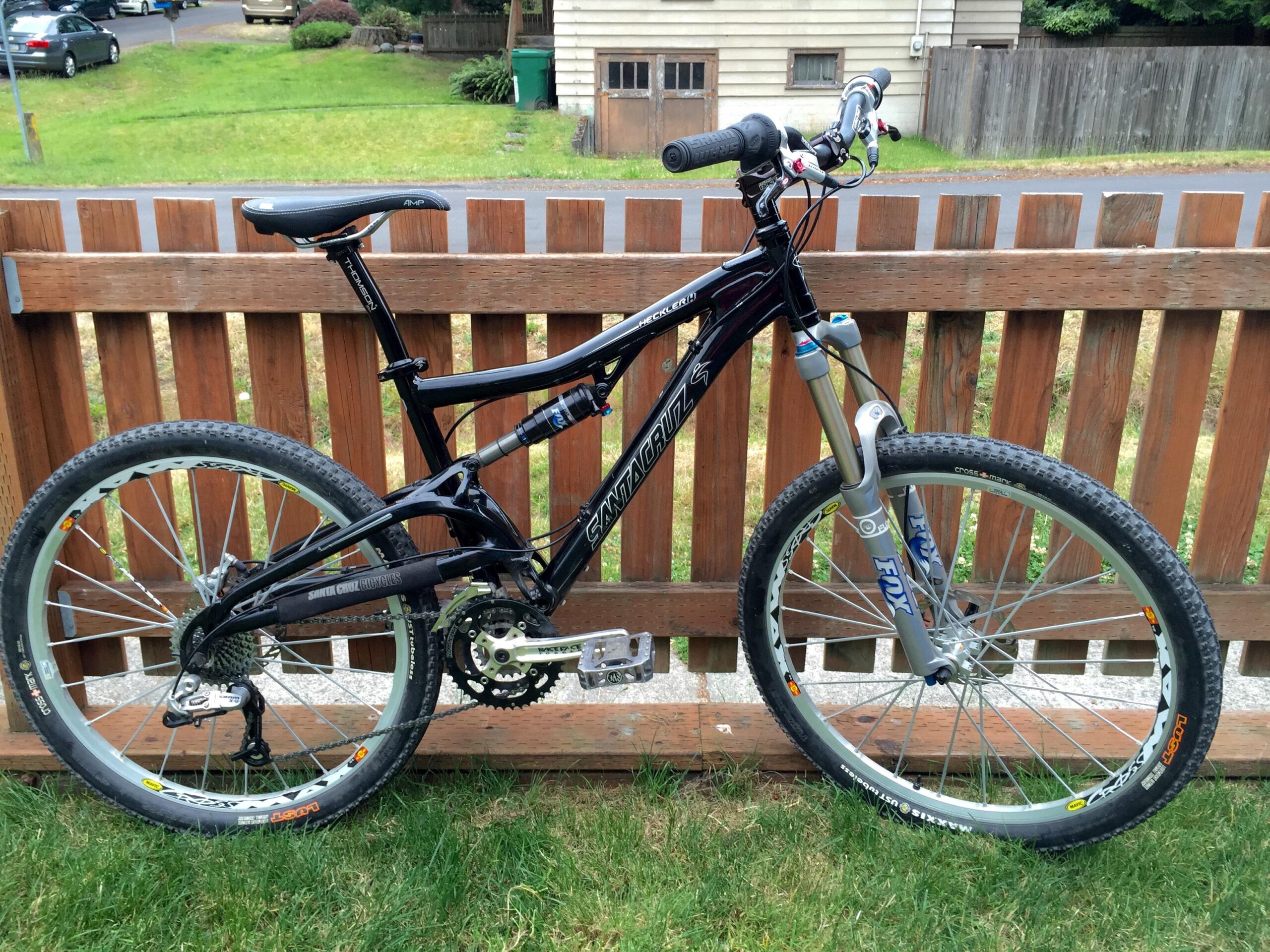 Santa Cruz Heckler: A black mountain bike with a prominent suspension system is positioned next to a wooden fence. The bike features wide tires with visible tread patterns and a sleek frame. The background shows a grassy area and a glimpse of a neighborhood street with parked cars.