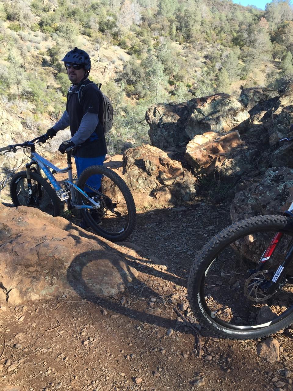 A person wearing a helmet and sunglasses stands next to a blue mountain bike on rocky terrain, with a second bike visible in the foreground. The background features a wooded hillside with trees and scattered rocks under clear blue skies. Hidden Falls Regional Park mountain bike trail.