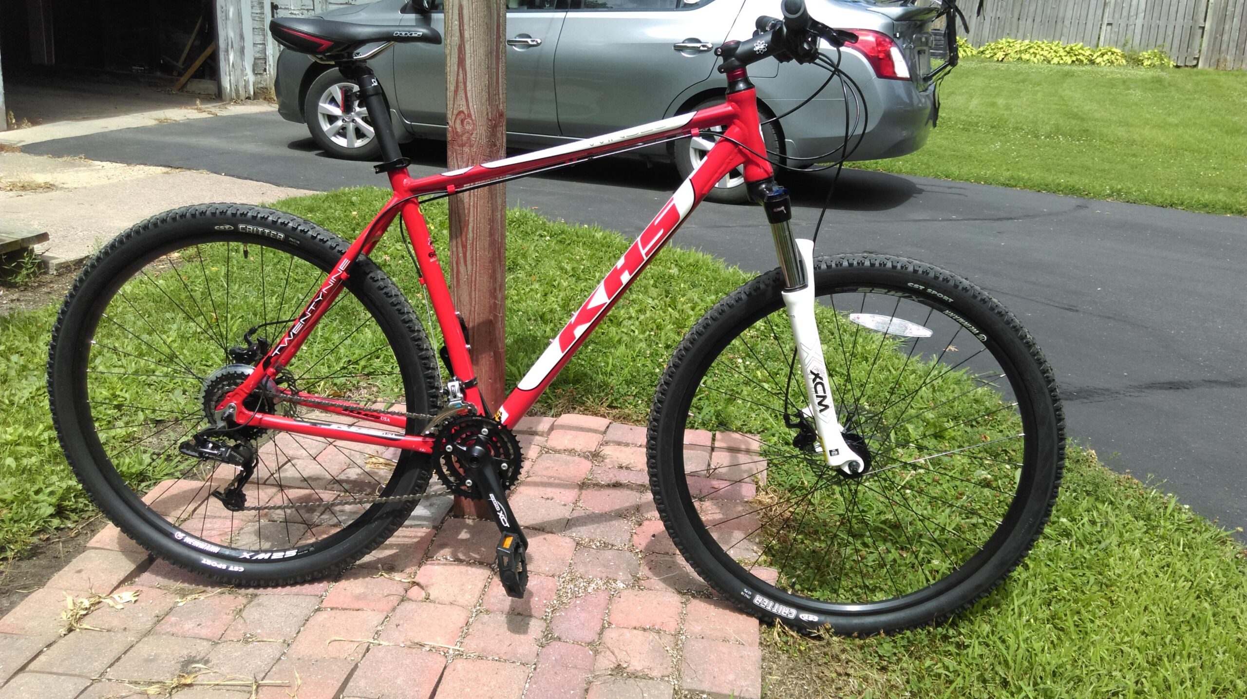 KHS Winslow: A red mountain bike with black and white accents is leaning against a wooden post on a brick pathway, set in a grassy area. In the background, a gray car is parked on a paved driveway, and there's a wooden fence and greenery visible further back.