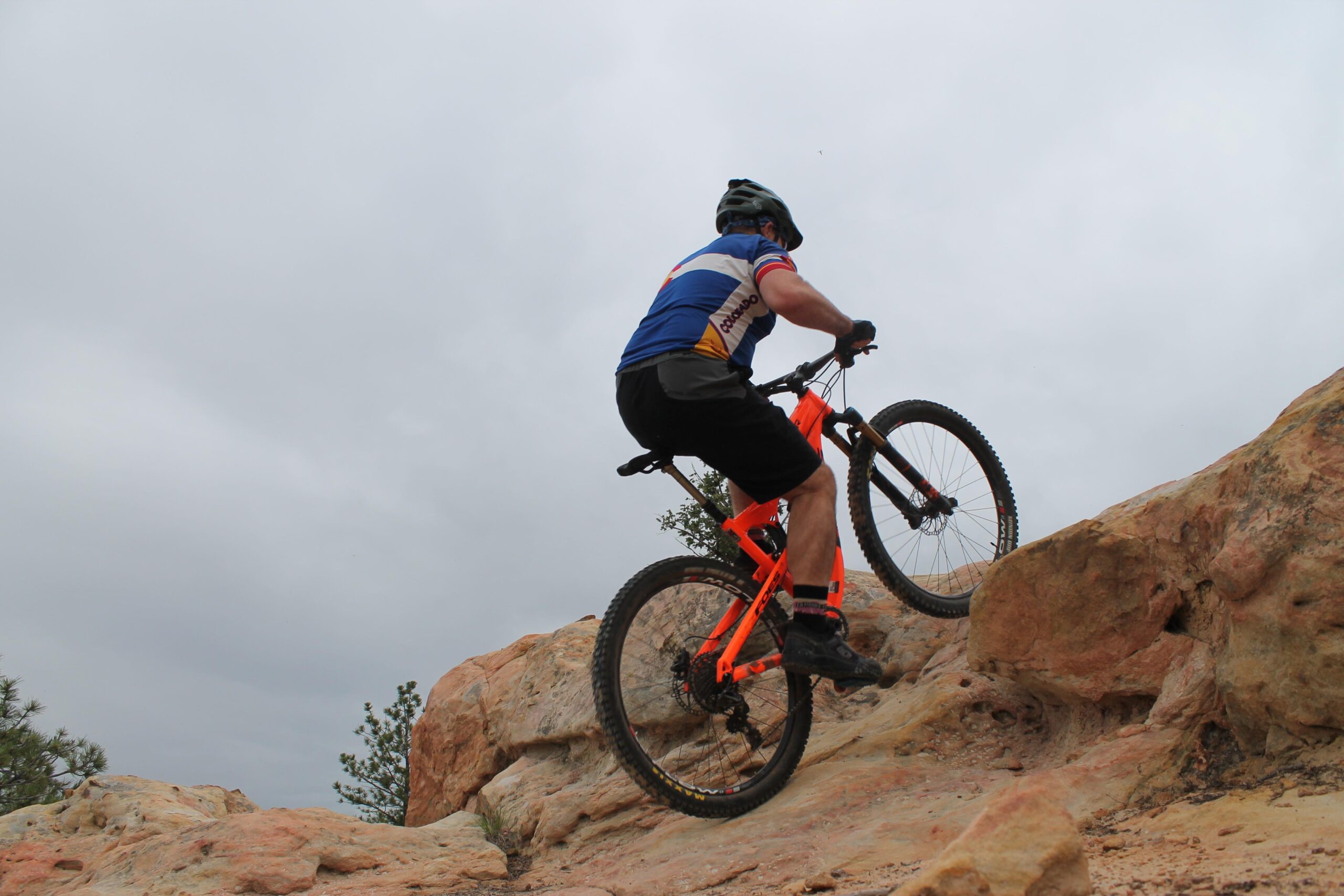 A mountain biker wearing a blue and white jersey and black shorts is riding an orange bike up a rocky slope on an overcast day. The scene features natural rocky terrain and tufts of greenery in the background. Ute Valley Park mountain bike trail.