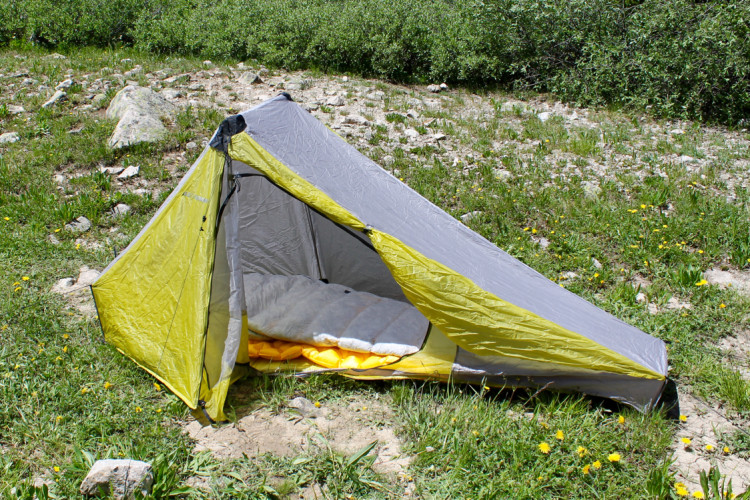 A lightweight, yellow and gray camping tent set up on a grassy area dotted with small rocks and wildflowers. The tent's entrance is open, revealing a sleeping pad inside. The surrounding landscape features green shrubs and a natural, outdoor environment.