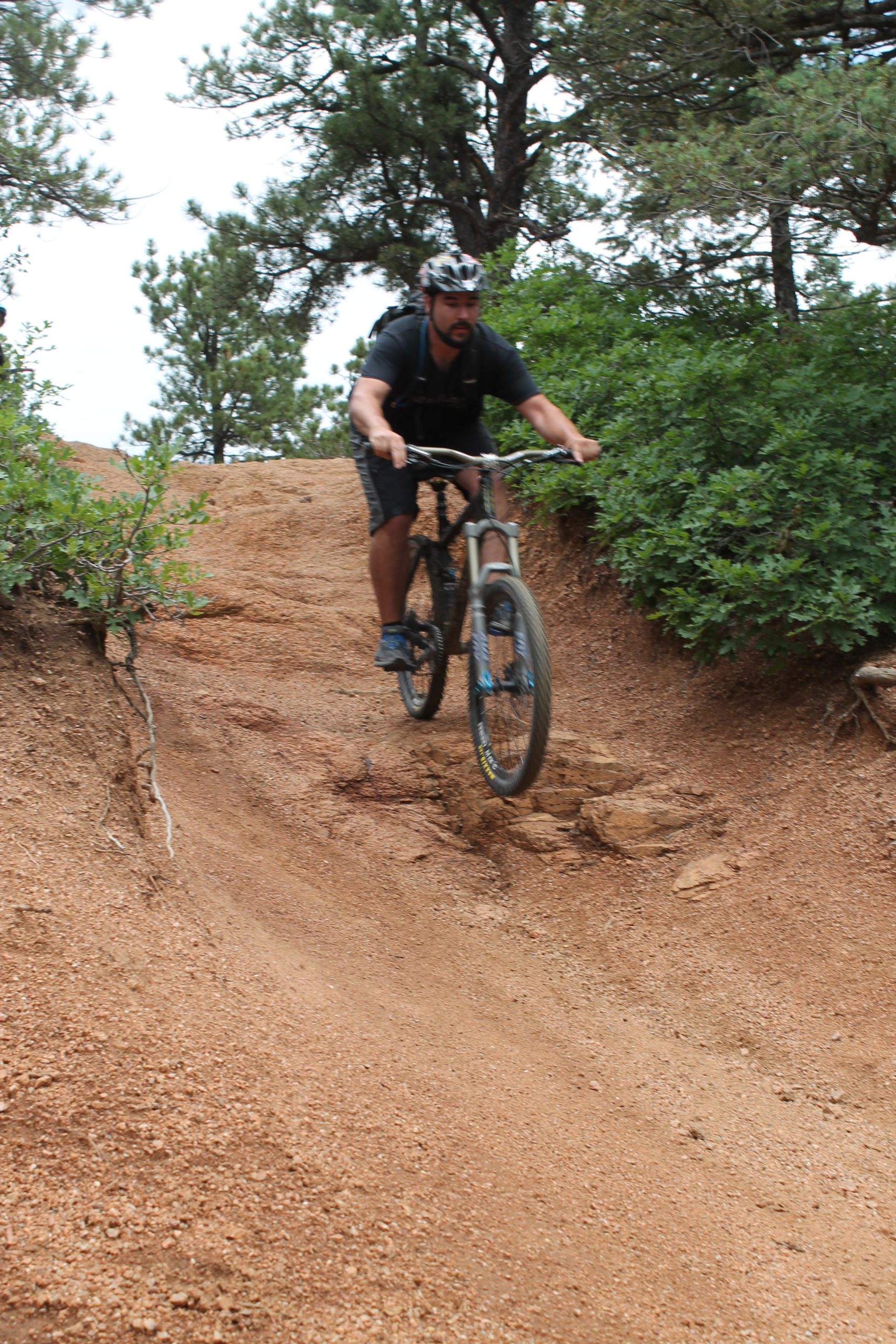 A mountain biker performing a jump on a dirt trail surrounded by pine trees and greenery. The rider is in mid-air, showcasing an action-packed moment on the trail. Captain Jack's mountain bike trail.