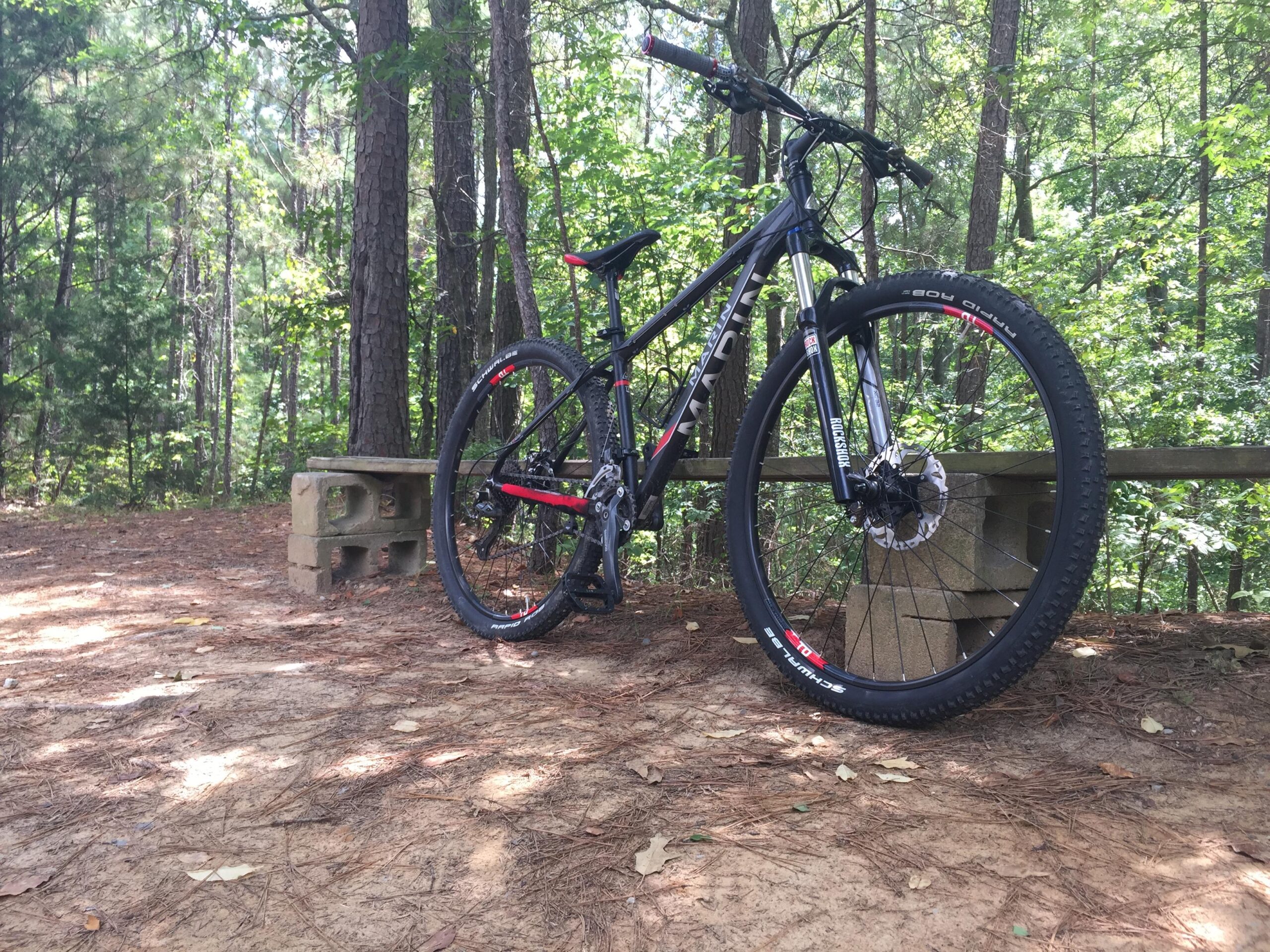 Marin Marin Bolinas Ridge hydro: A black and red mountain bike is resting on a sandy area next to a stone bench, surrounded by tall green trees in a wooded setting.