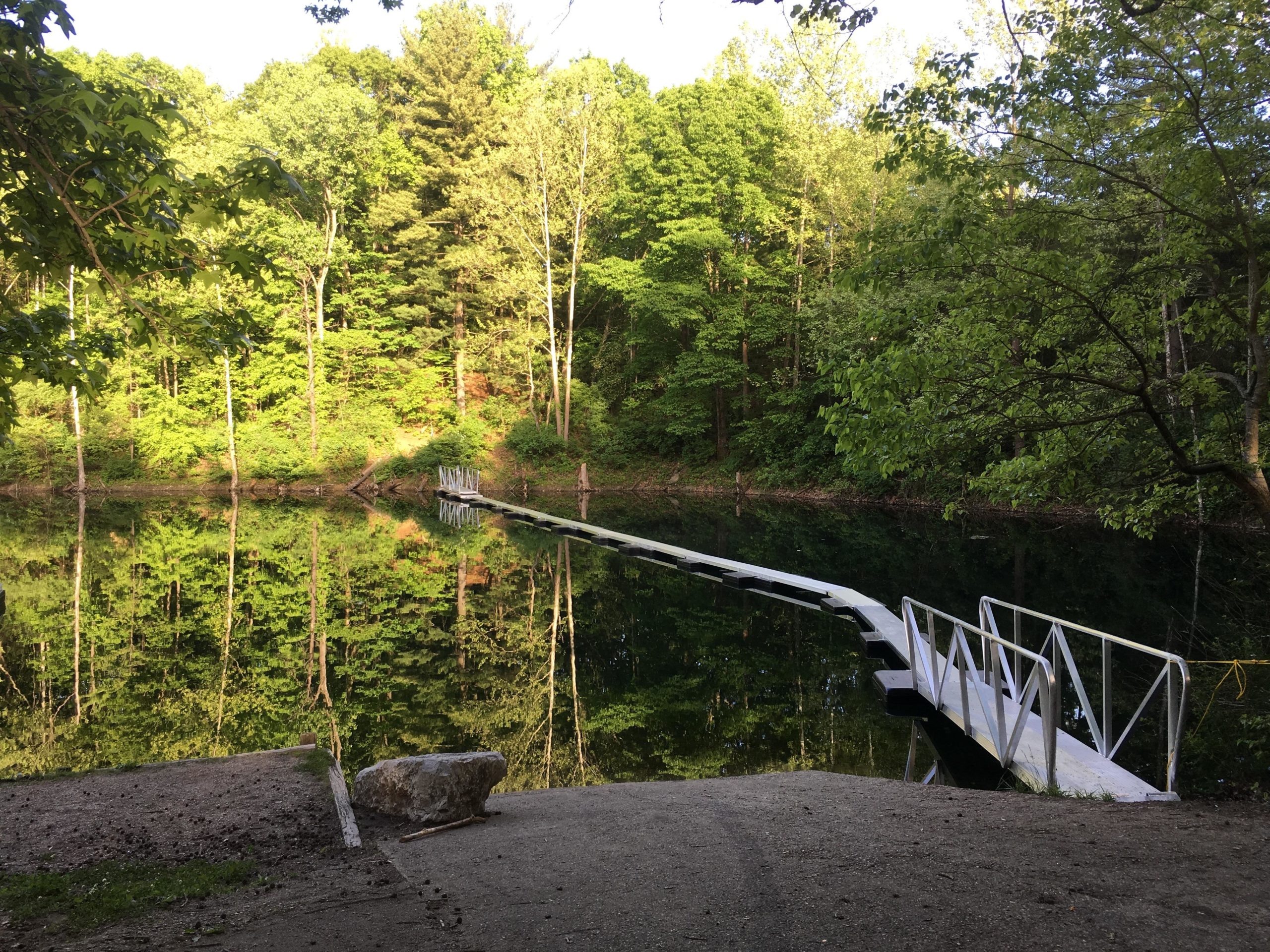 A serene forest scene featuring a wooden walkway extending over a calm lake, surrounded by lush green trees. The water reflects the vibrant foliage, creating a peaceful and natural atmosphere. Sunlight filters through the leaves, illuminating the path and water. Floating Water Trail mountain bike trail.