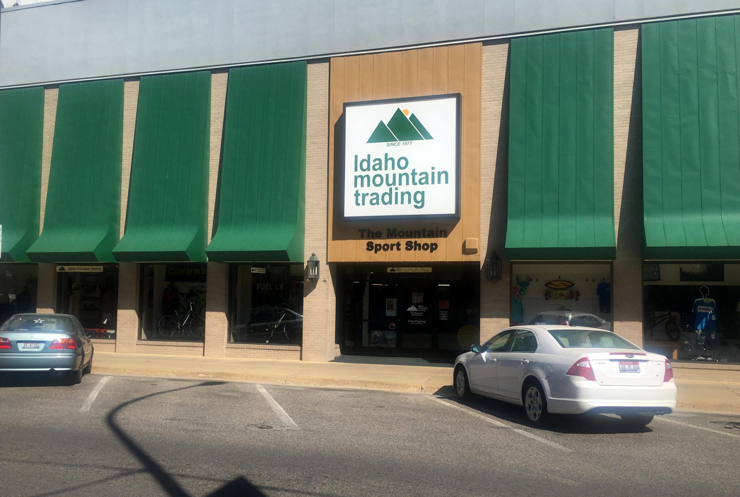 Storefront of Idaho Mountain Trading, featuring green awnings and a large sign that reads "Idaho Mountain Trading" and "The Mountain Sport Shop." The windows display bicycles and outdoor gear. Parked cars are visible in front of the store.