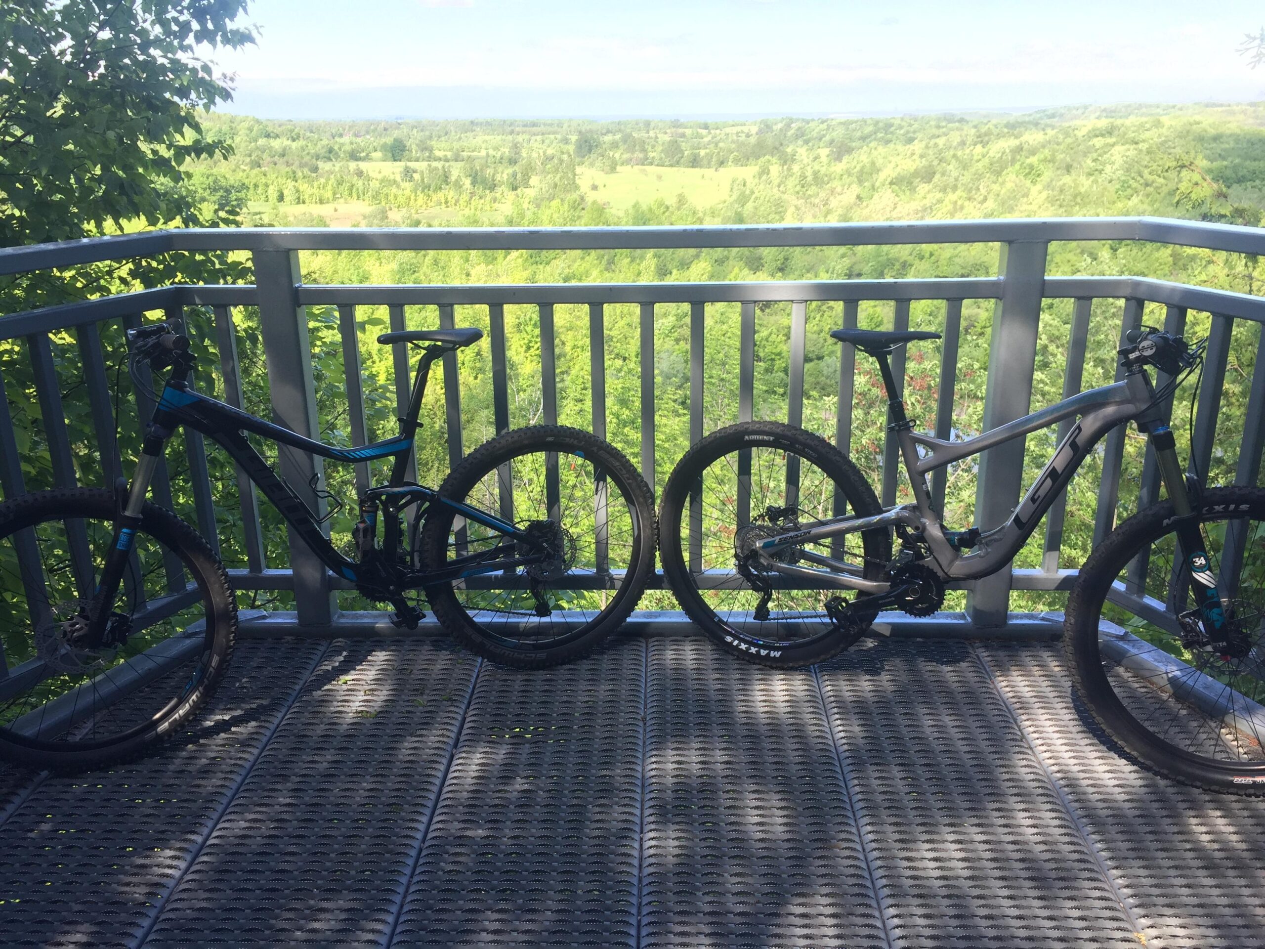 Giant Trance 27.5 3: Two mountain bikes, one blue and black and the other silver and black, are parked on a lookout deck surrounded by lush green trees. The deck offers a scenic view of the landscape in the background.