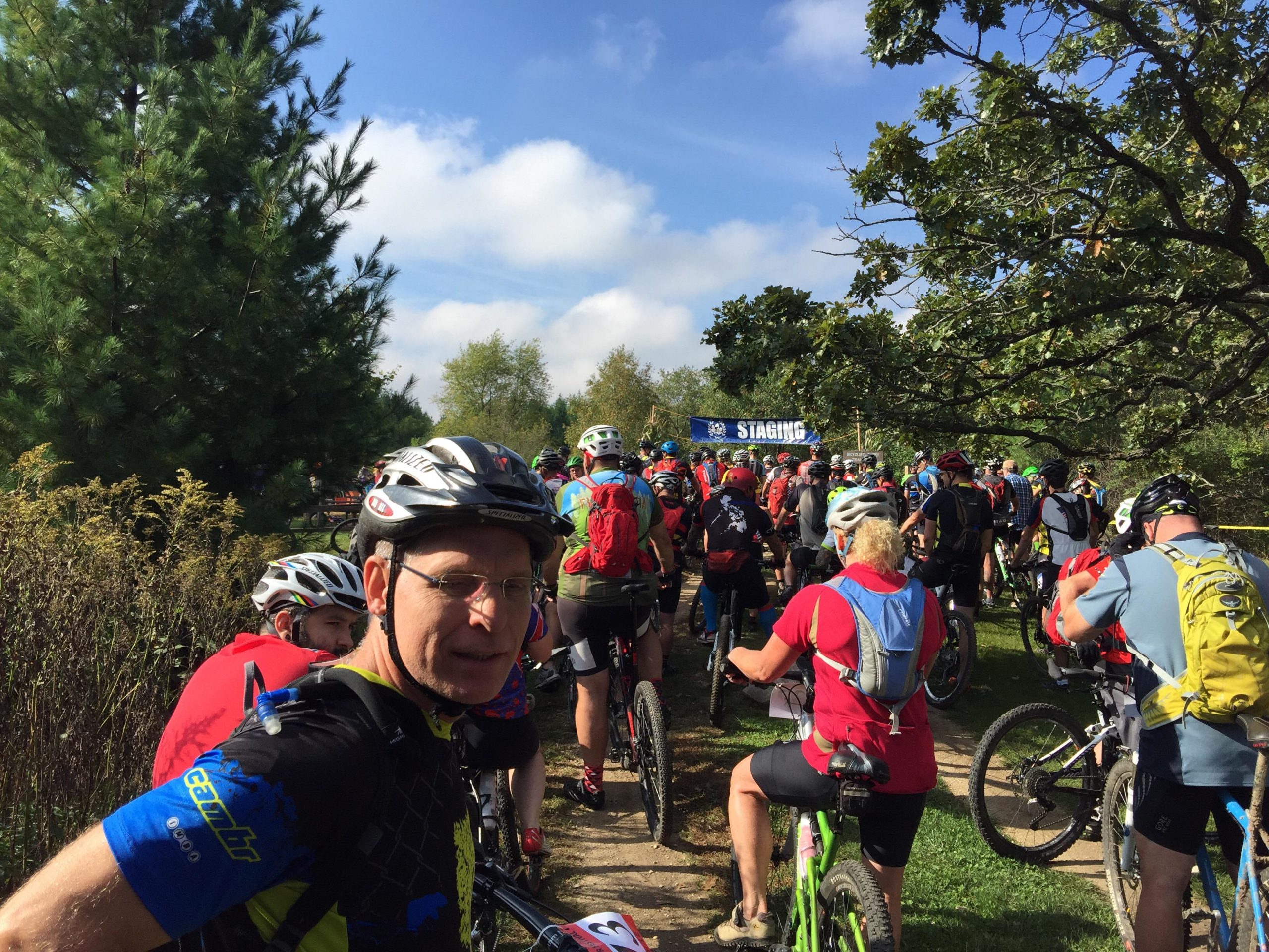 A group of mountain bikers lining up at the starting point of an event, surrounded by trees and greenery under a partly cloudy sky. The scene includes cyclists wearing helmets and various biking gear, with a "STAGING" banner visible in the background. John Muir Trails mountain bike trail.