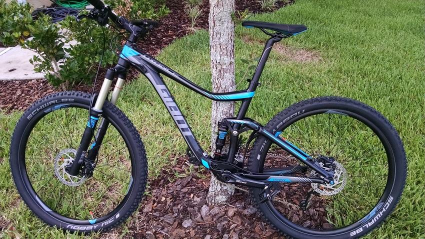 Giant Trance 27.5 3: A modern mountain bike with a black and blue frame is leaning against a tree. The bike features thick tires suitable for off-road riding, a front suspension fork, and a sleek design. The surrounding area has green grass and mulch, indicating a well-maintained outdoor space.