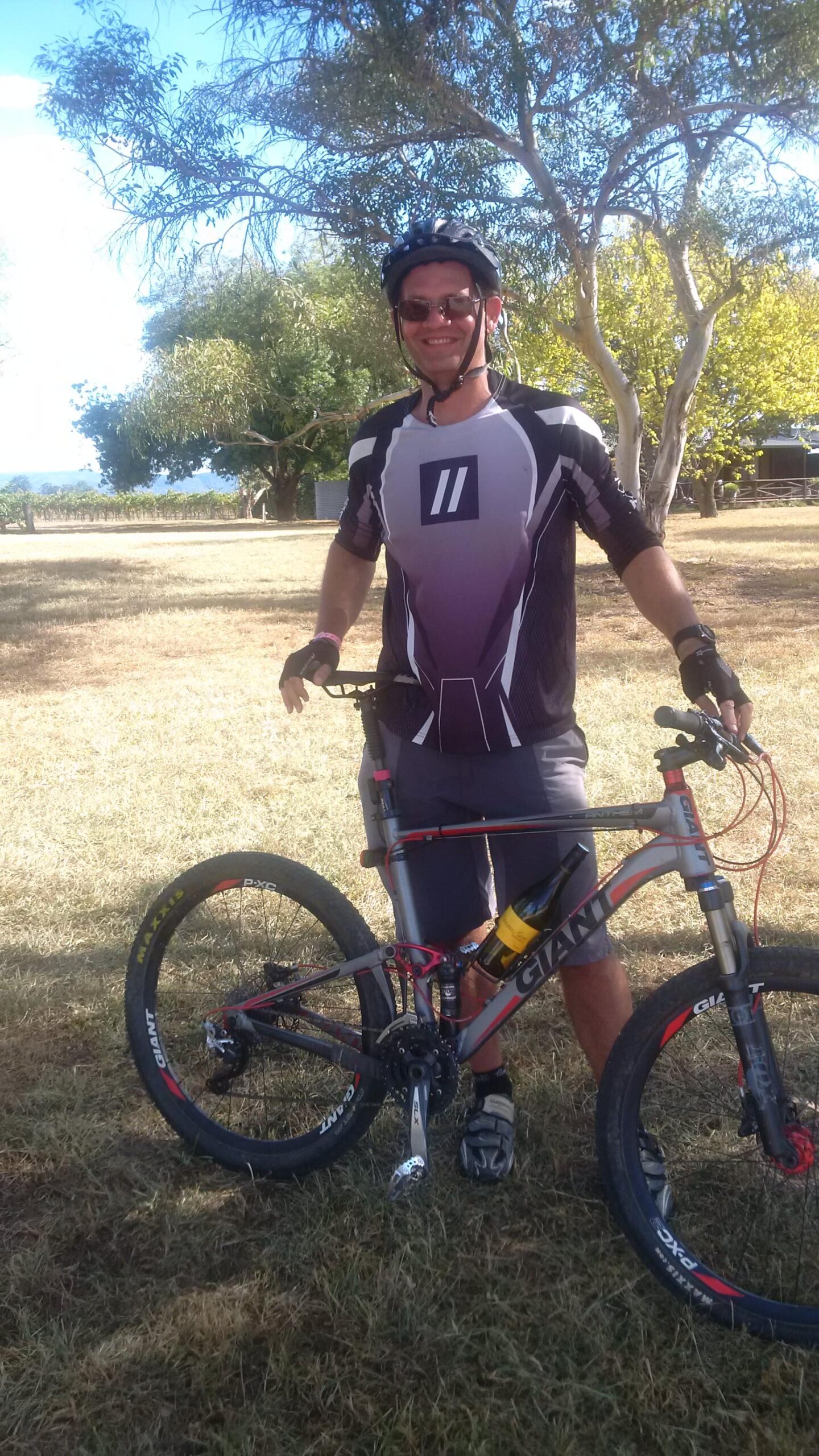 Giant Anthem: A smiling person wearing a cycling helmet and jersey stands next to a mountain bike on grassy terrain. The bike has a bottle of wine attached to it, and trees and a blue sky are visible in the background.