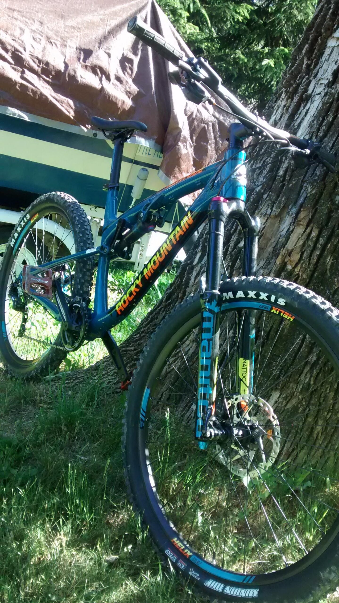 Rocky Mountain Altitude: A blue and orange Rocky Mountain mountain bike leaning against a tree in a grassy area. The bike features thick tires with "Maxxis" branding and visible suspension components. A covered boat is partially visible in the background, with trees providing shade above.