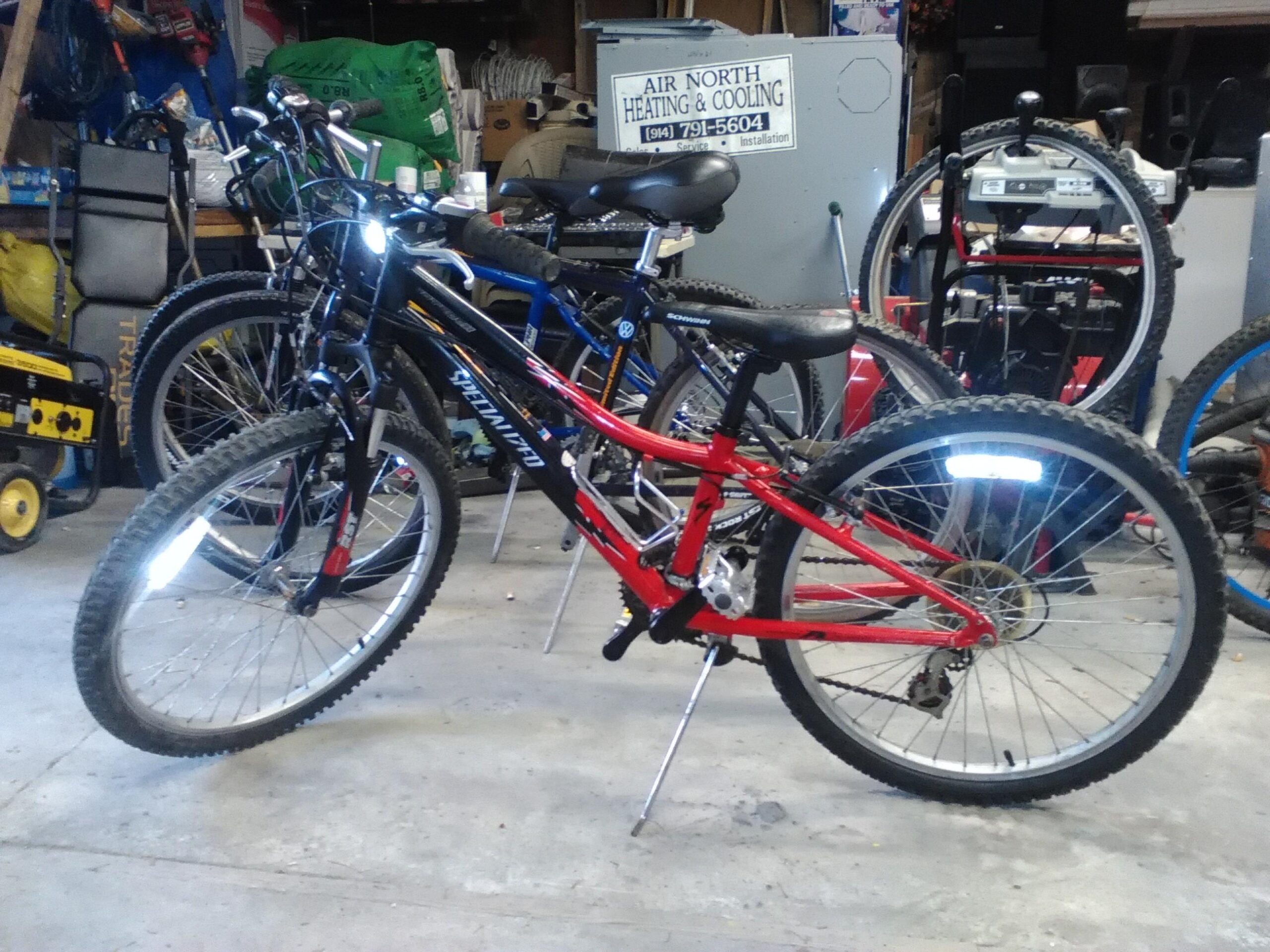 Alt text: A red and black mountain bike with a kickstand, positioned in a garage among other bikes. The garage has various tools and equipment in the background, indicating a cluttered workspace.