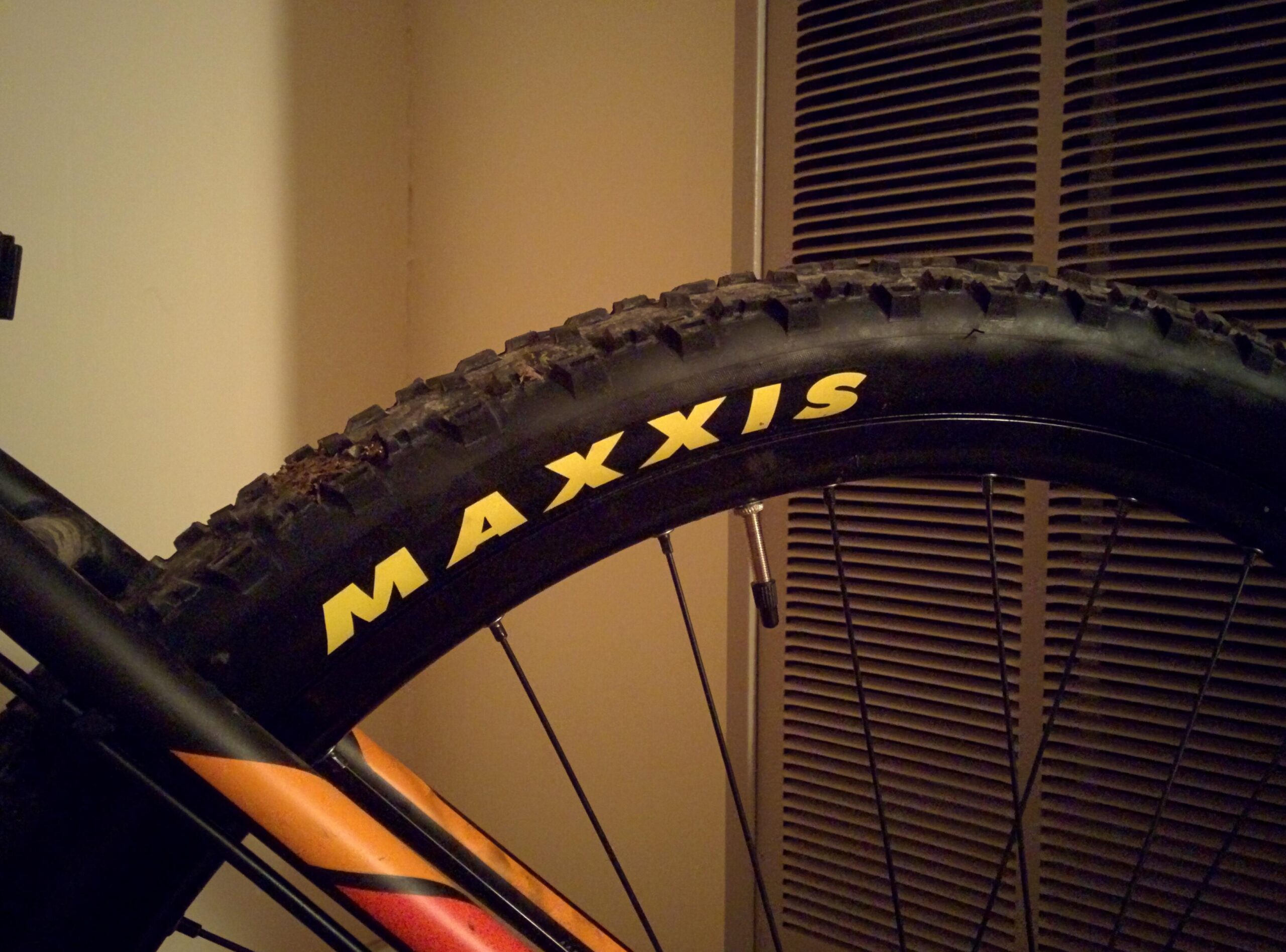 Trek Marlin 5 29er: A close-up view of a bicycle tire featuring the brand name "MAXXIS" in yellow letters. The tire has a textured surface designed for grip, and part of the bike's frame is visible alongside it, with an air valve showing. The background includes a neutral-colored wall and a vent.