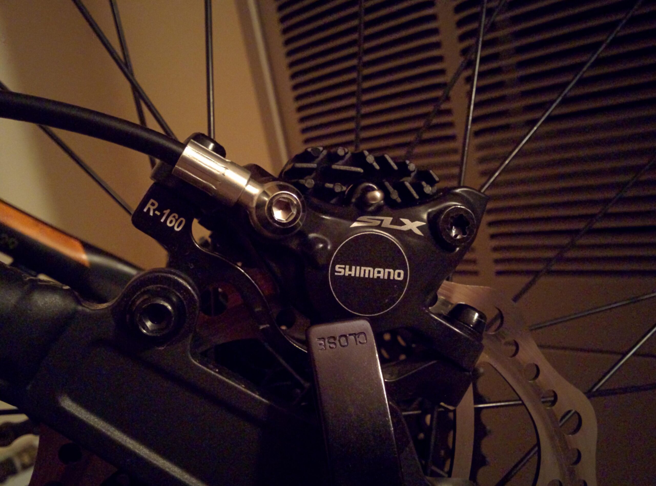 Trek Marlin 5 29er: Close-up image of a Shimano SLX bicycle brake caliper and rotor, featuring a black metallic finish. The brake line is partially visible, with a connector labeled "R-160." The image captures the intricate details of the brake assembly.