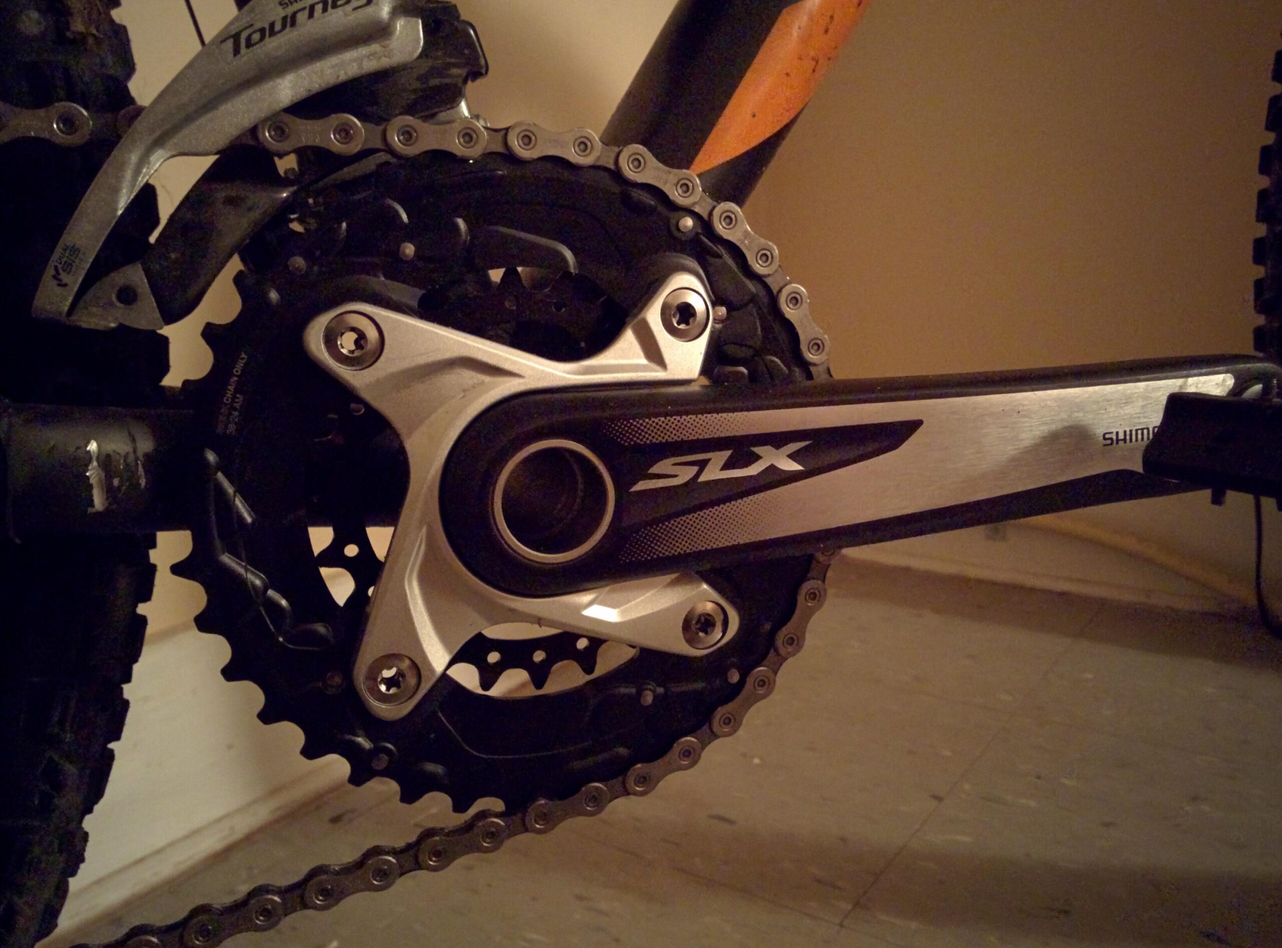 Trek Marlin 5 29er: Close-up of a bicycle crankset featuring Shimano SLX components, with a black chain and chainrings. The background is softly illuminated, highlighting the details of the crank and the bike's frame.