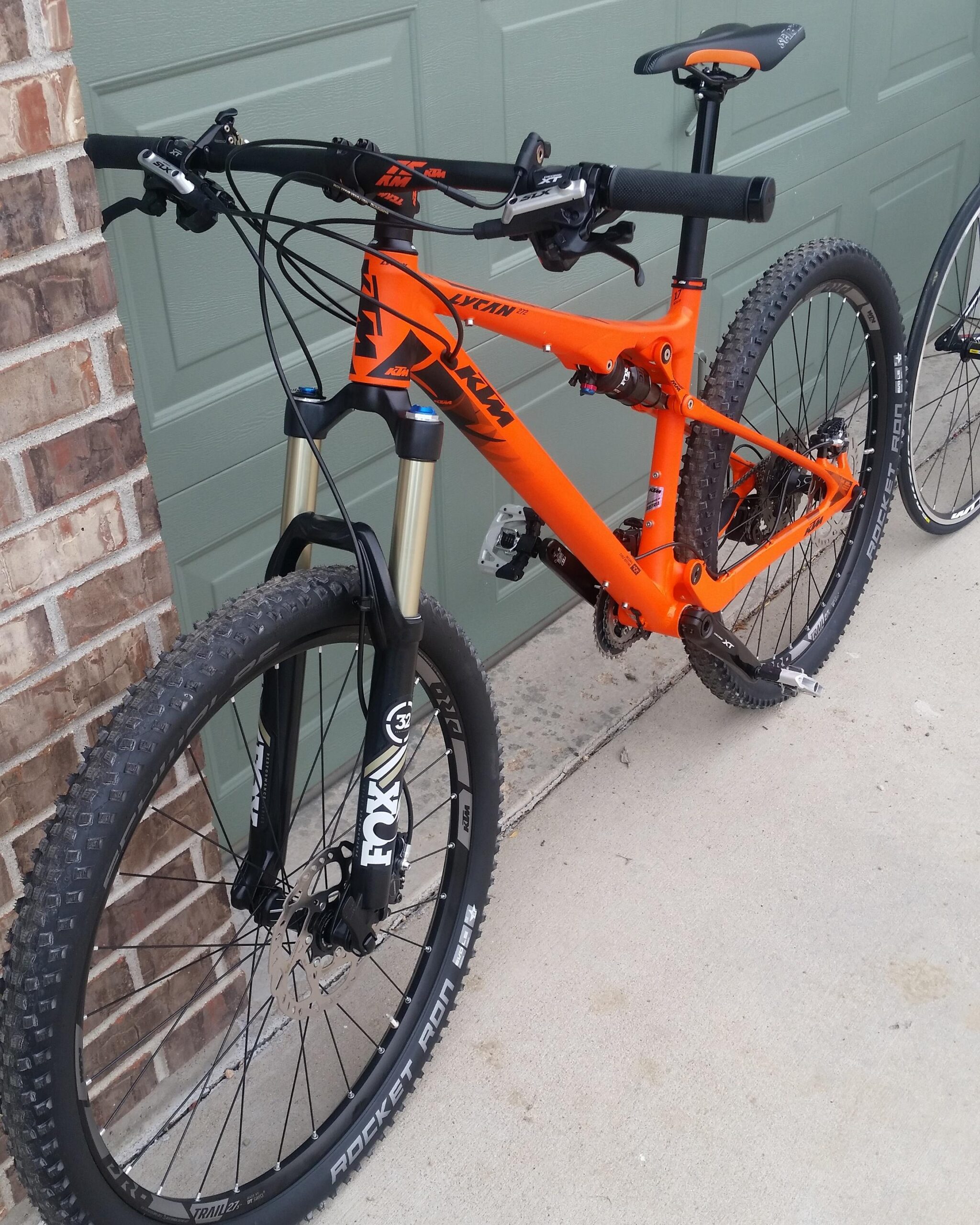 Ktm Lycan 272: A bright orange mountain bike with a sleek design, featuring front suspension forks, knobby tires, and an ergonomic handlebar setup. The bike is positioned near a garage door, highlighting its vibrant color against the muted background.