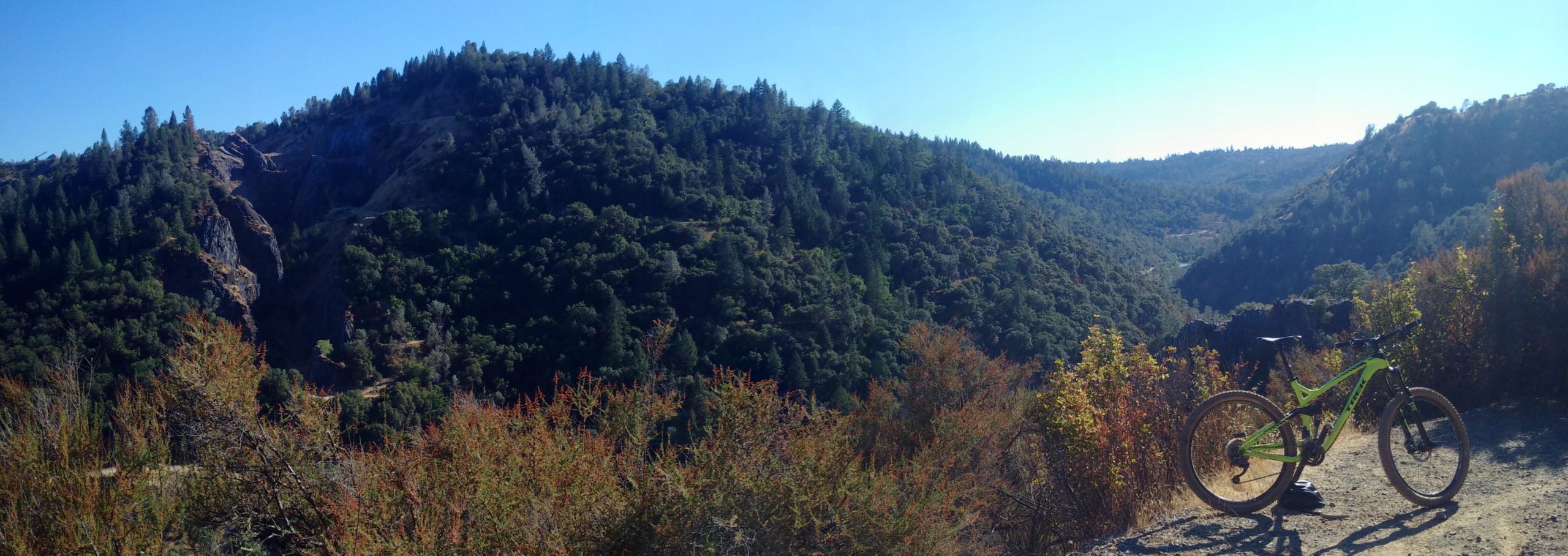 A panoramic view of a lush green landscape featuring rolling hills and dense forests under a clear blue sky, with a mountain bike positioned in the foreground on a dirt path. The scene captures the essence of outdoor adventure and natural beauty. Stagecoach / Flood / Manzanita mountain bike trail.