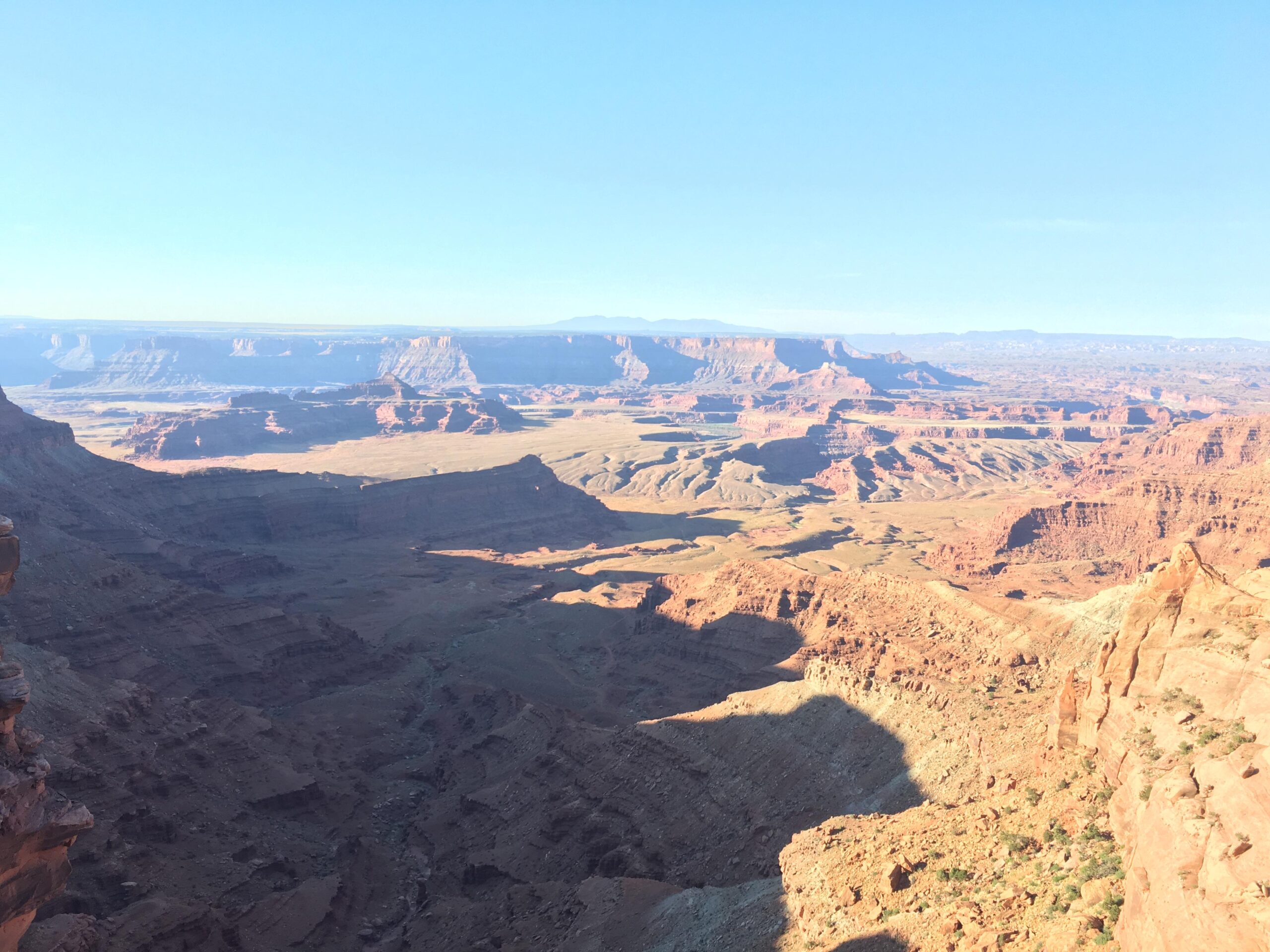 A panoramic view of a rugged canyon landscape featuring vibrant red rock formations, layered canyons, and distant mesas under a clear blue sky. Shadows create depth in the terrain, showcasing the natural beauty and vastness of the environment. Dead Horse Point State Park mountain bike trail.