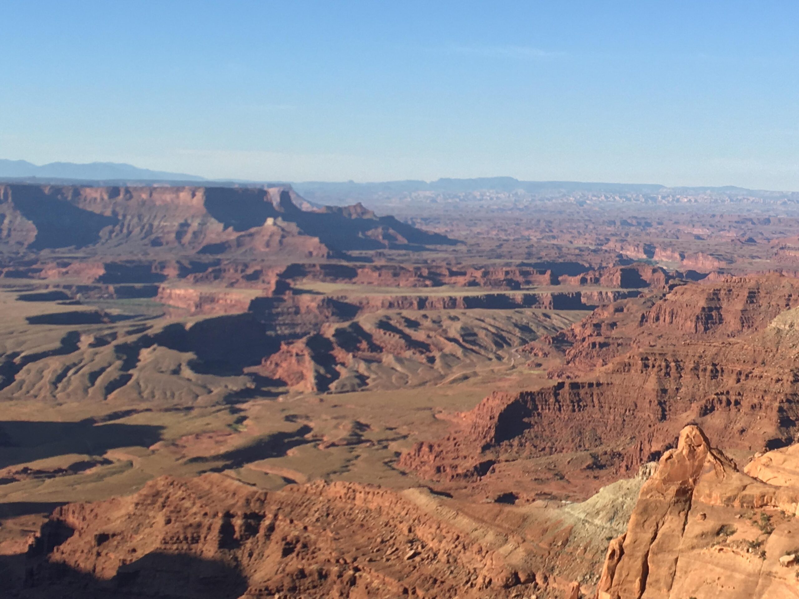 A vast, arid landscape featuring layered rock formations and rugged cliffs, under a clear blue sky. The terrain showcases a mix of red and brown hues, with shadows creating depth across the valleys and hills. Dead Horse Point State Park mountain bike trail.