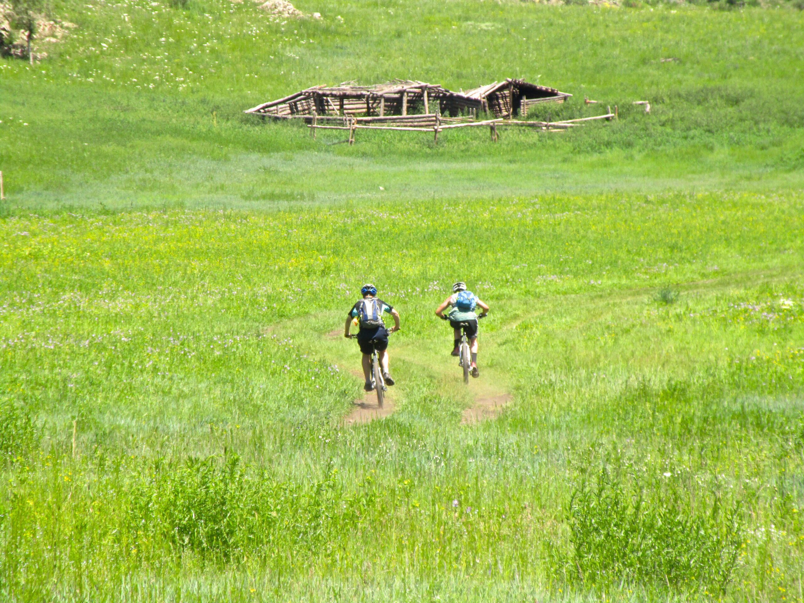 Two mountain bikers riding on a dirt path through a vibrant green meadow, with a rustic wooden structure in the background under a clear blue sky. Khargana Gol mountain bike trail.