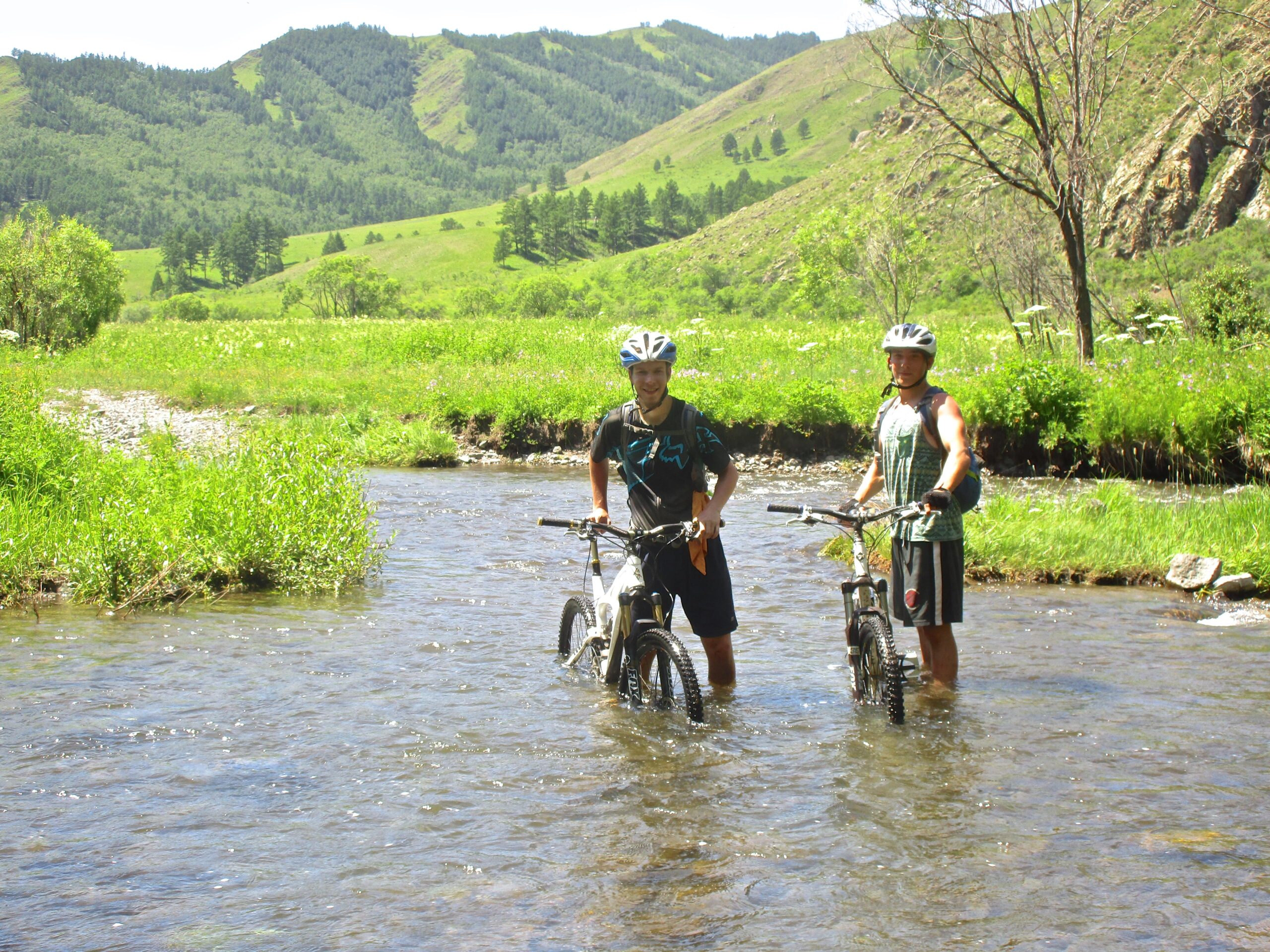 Two mountain bikers stand in shallow water, holding their bikes, amidst a lush green landscape with rolling hills and trees in the background on a sunny day. Khargana Gol mountain bike trail.