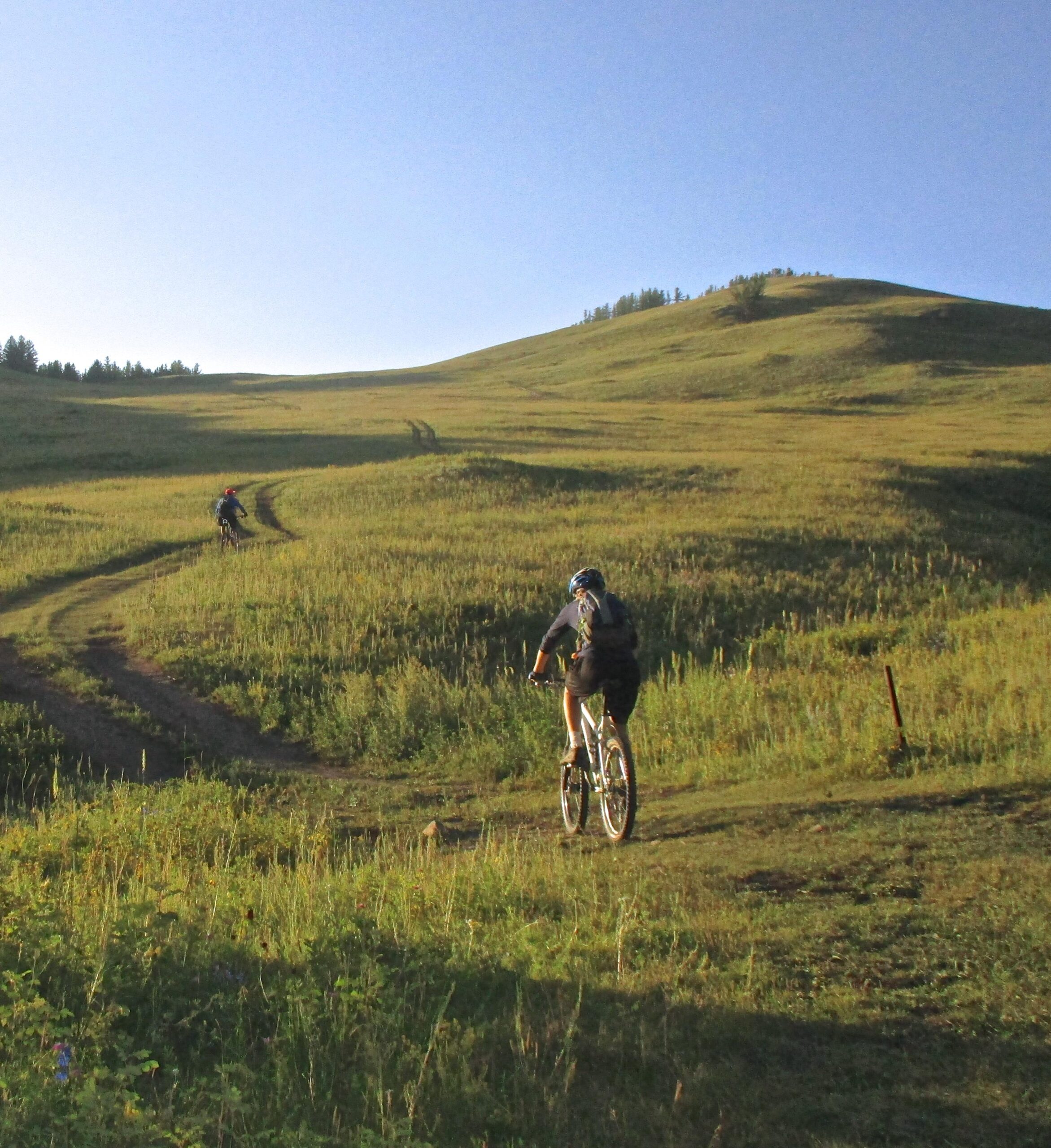 Two mountain bikers riding along a winding dirt path through a grassy landscape under a clear blue sky. Hills and trees are visible in the background, suggesting a serene outdoor setting. Selkh Am mountain bike trail.