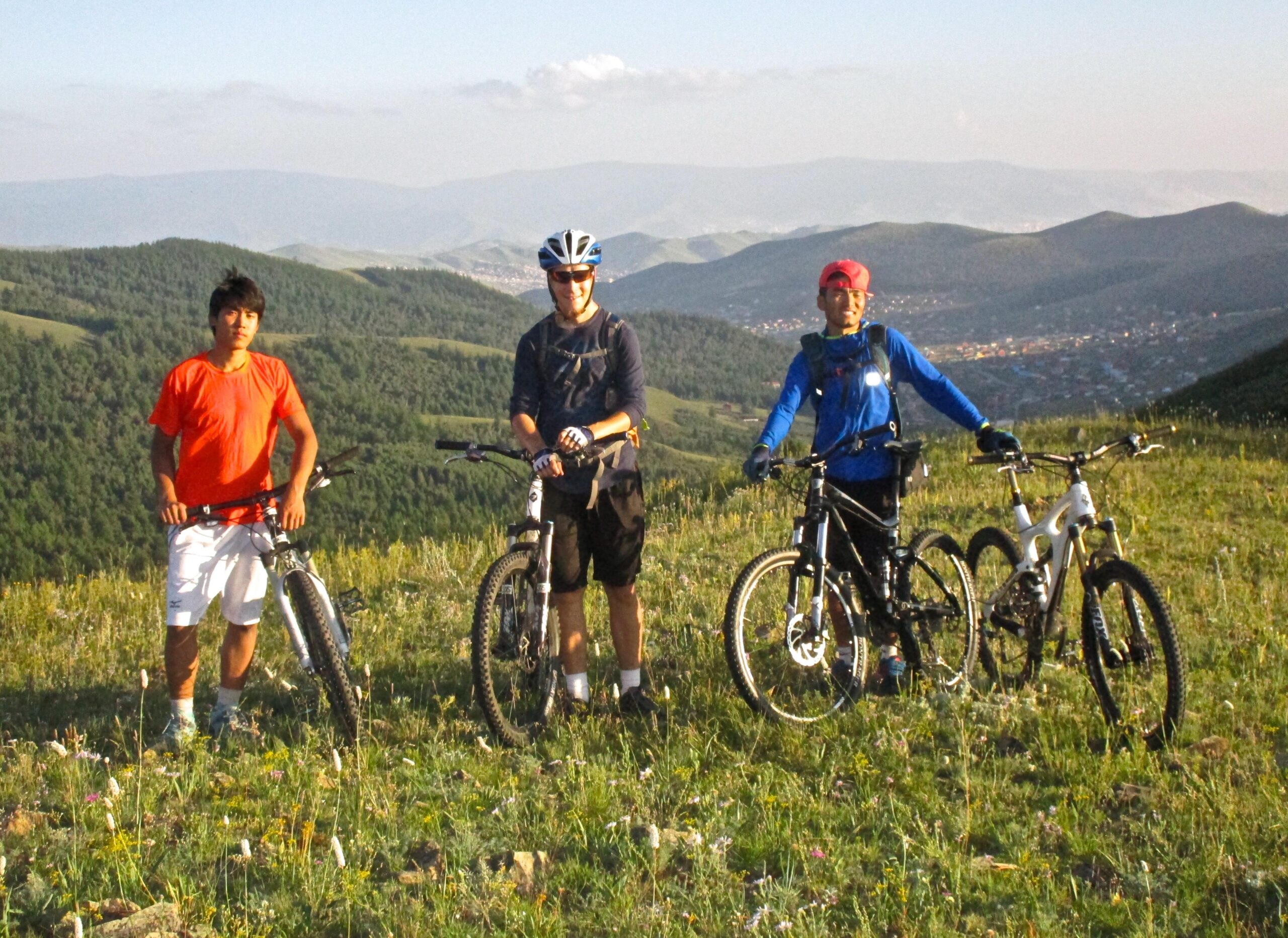 Three mountain bikers standing on a grassy hillside with a scenic view of rolling hills and a valley in the background. They are posed with their bikes, wearing casual athletic clothing. The atmosphere appears sunny and vibrant, highlighting the outdoorsy adventure spirit. Selkh Am mountain bike trail.