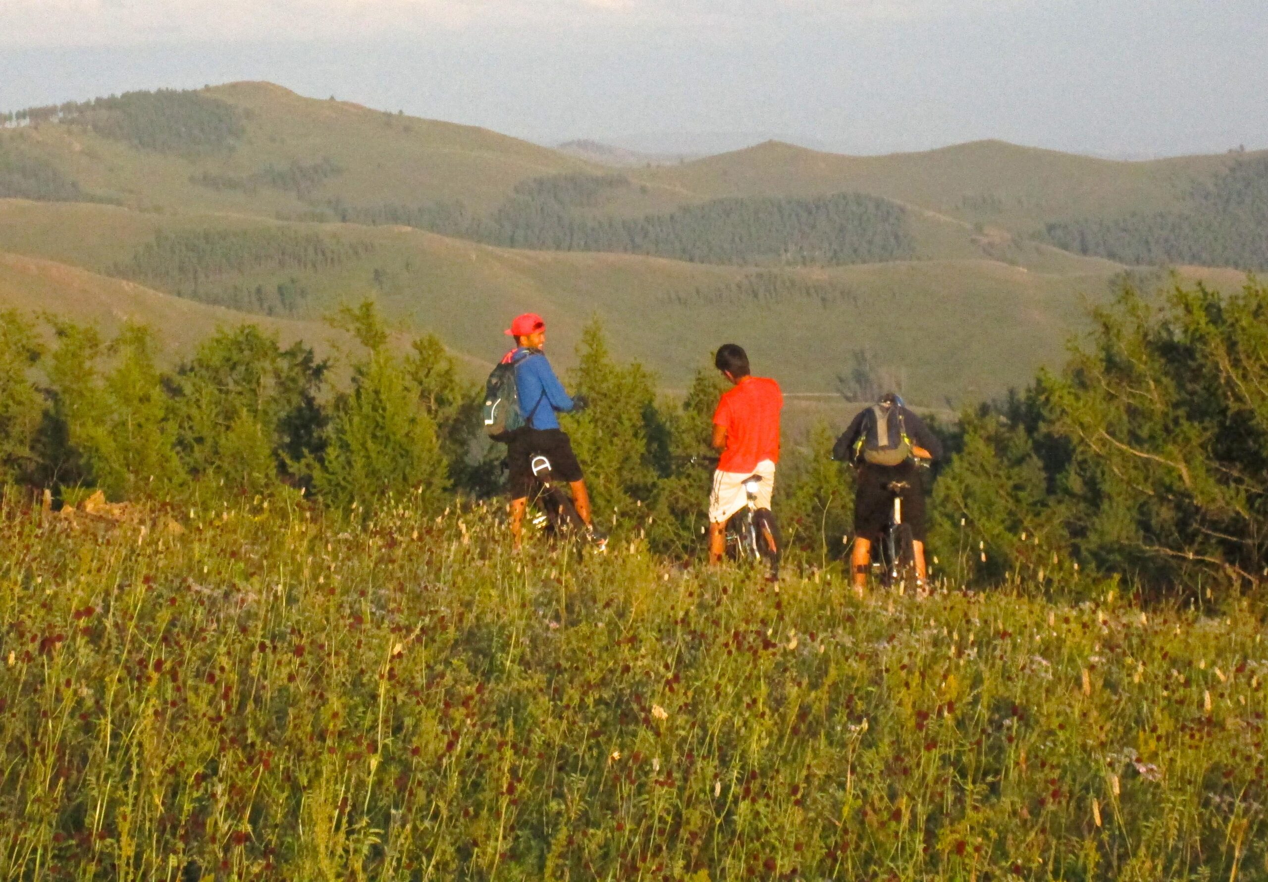Three individuals are standing on a grassy hill covered in wildflowers, overlooking a landscape of rolling green hills and trees. One person is wearing a red hat and a blue jacket, another is dressed in a red shirt, and the third is in dark clothing. They appear to be enjoying a moment of pause during an outdoor activity, possibly mountain biking or hiking. Selkh Am mountain bike trail.